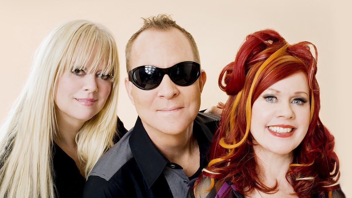 Three people posing together, one with long blonde hair, one wearing black sunglasses, and one with red and orange styled hair, all smiling.