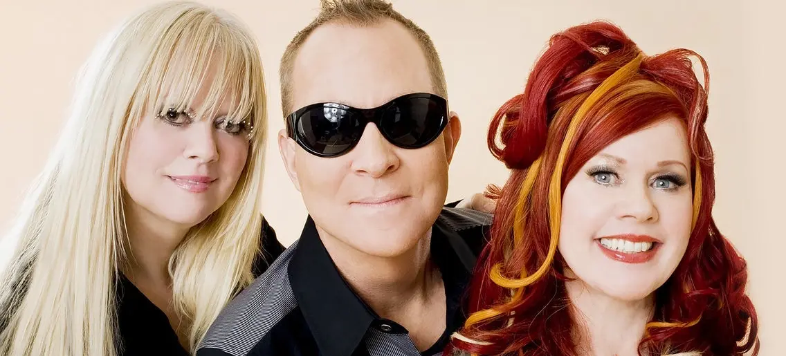 Three people posing together, one with long blonde hair, one wearing black sunglasses, and one with red and orange styled hair, all smiling.