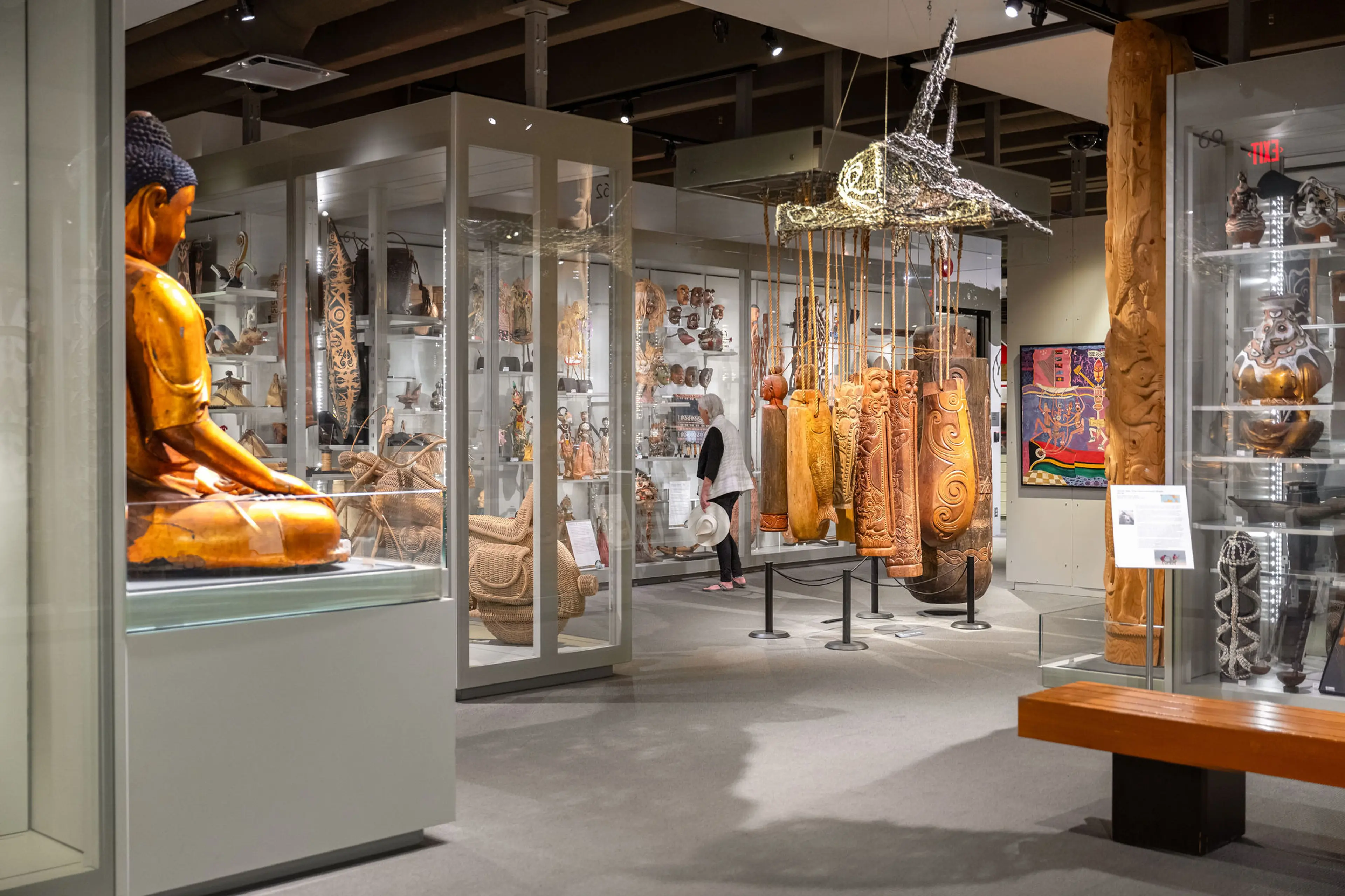 Museum gallery with wooden sculptures, hanging carved wooden panels, and a person viewing exhibits.