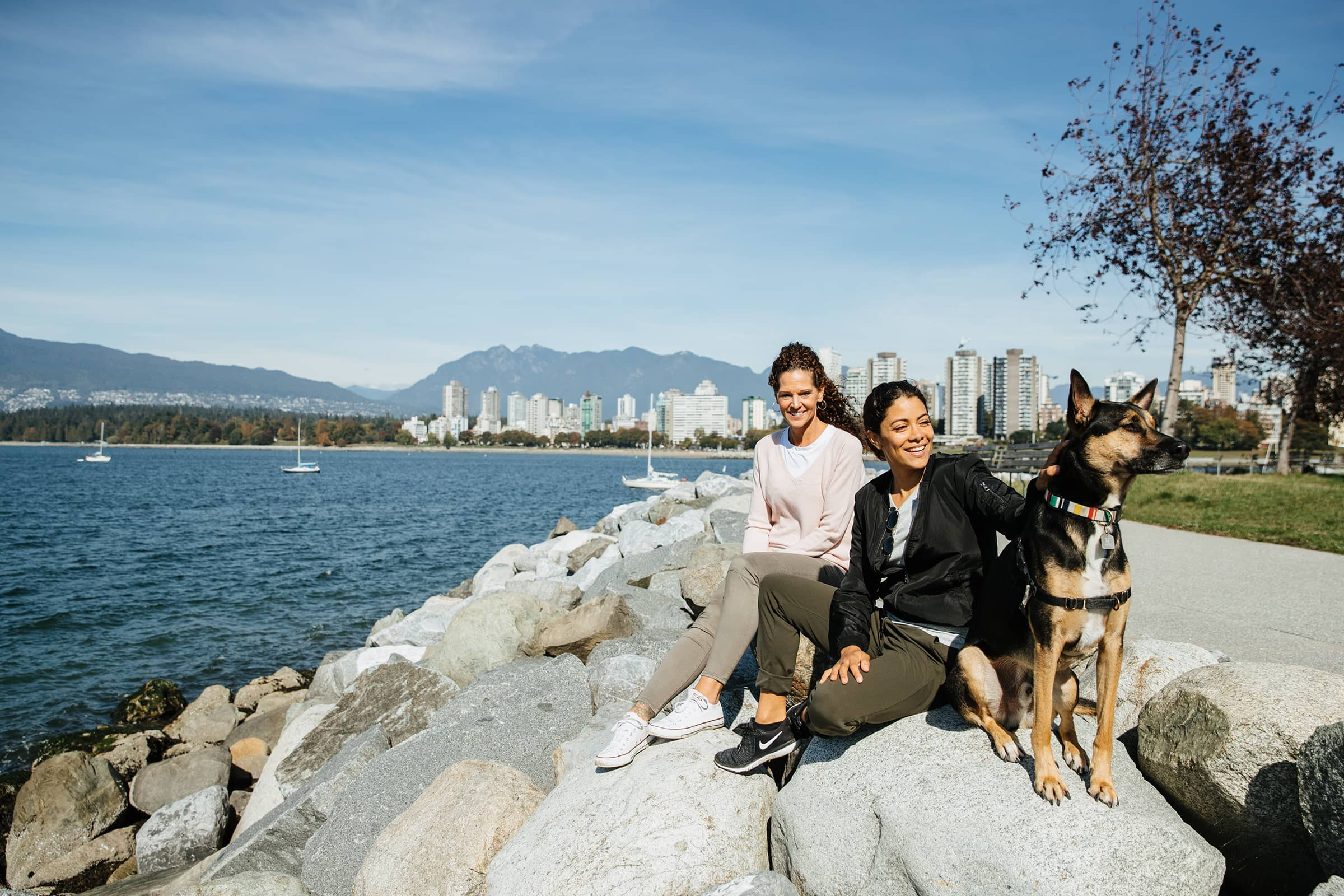 Two women sitting on rocks by the water with a dog, city skyline and mountains in the background.