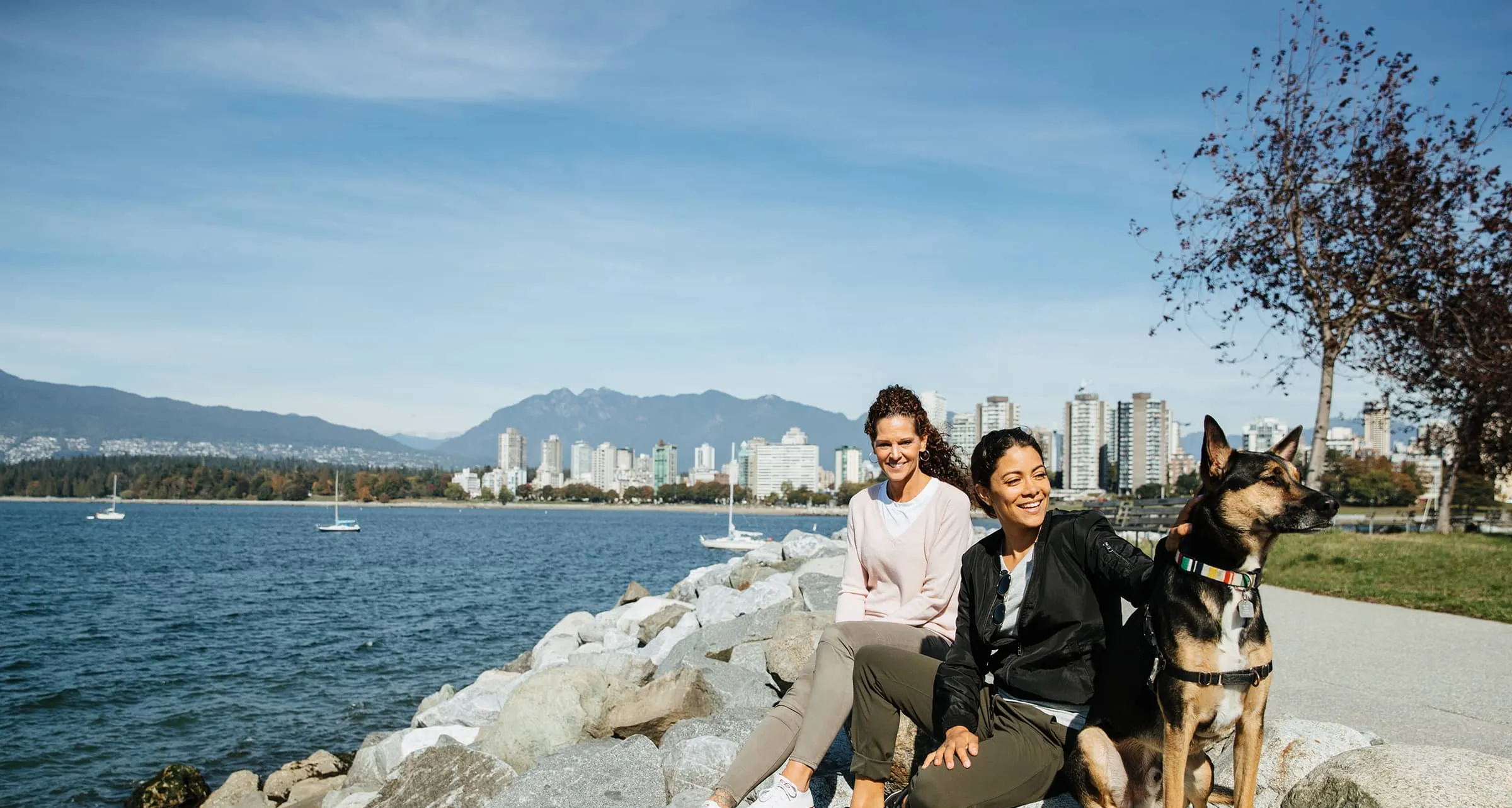 Two women sitting on rocks by the water with a dog, city skyline and mountains in the background.