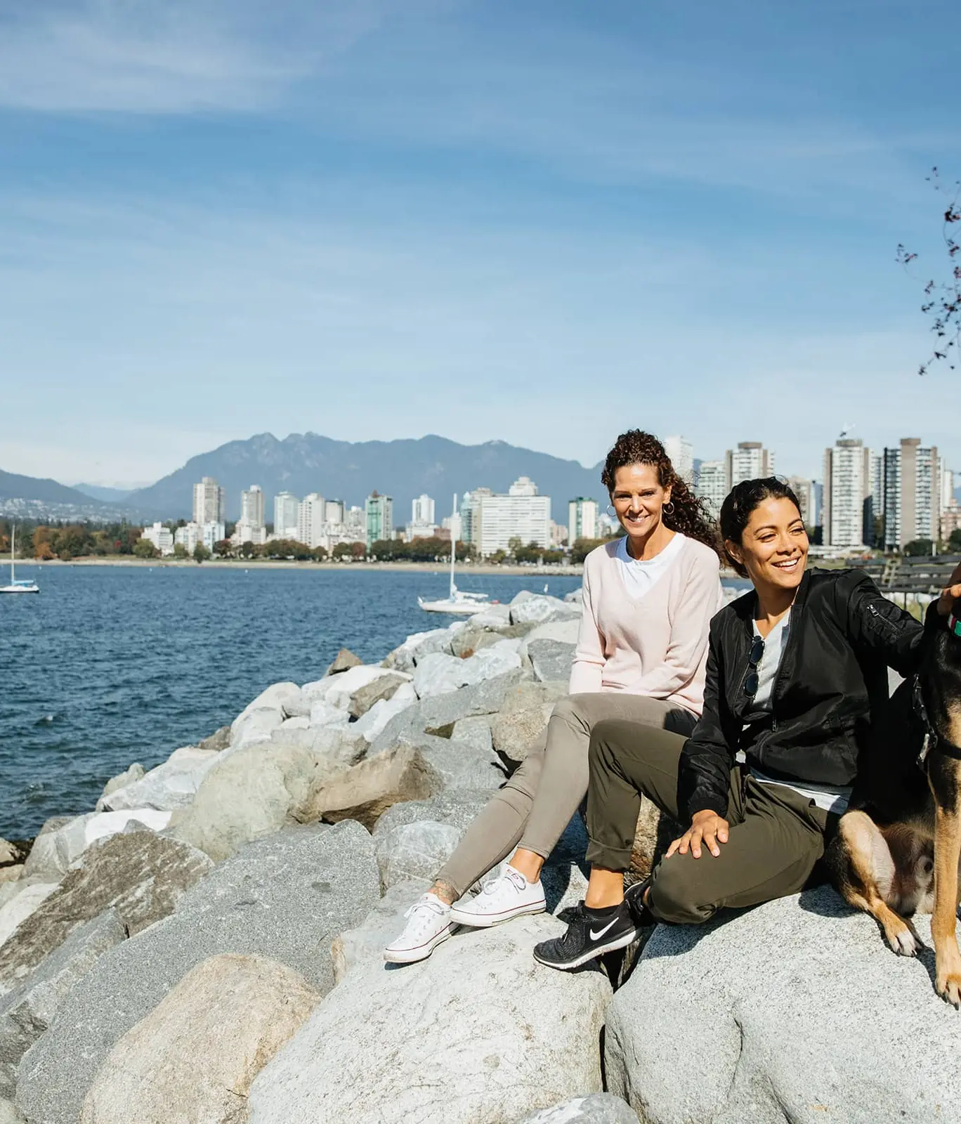 Two women sitting on rocks by the water with a dog, city skyline and mountains in the background.