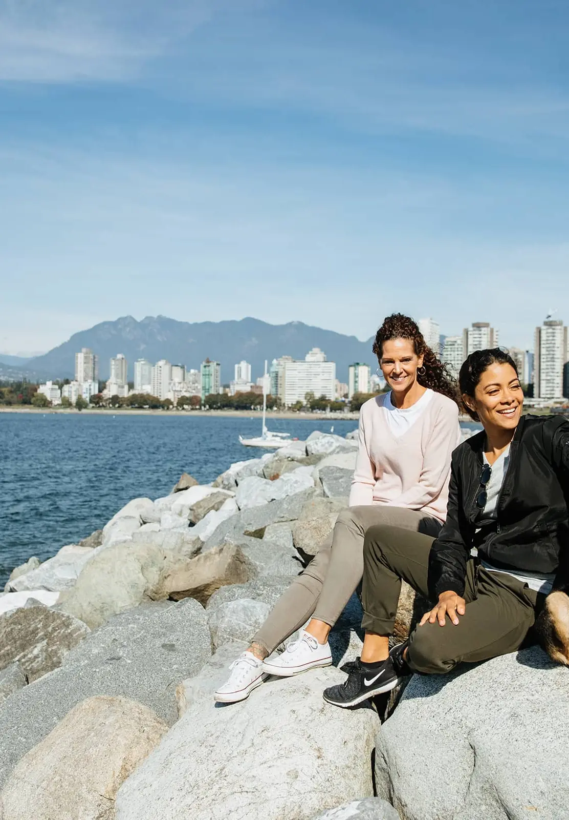 Two women sitting on rocks by the water with a dog, city skyline and mountains in the background.