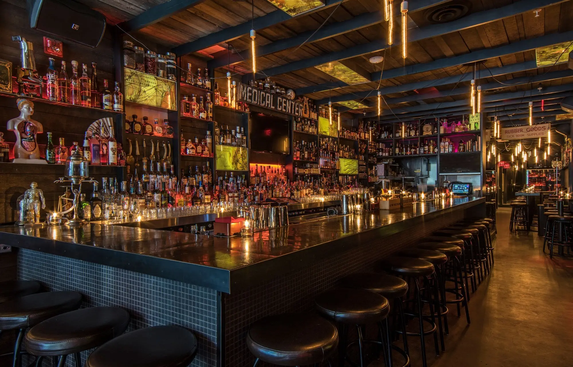 Long bar counter with many stools and shelves filled with bottles and decorations in a dimly lit bar.