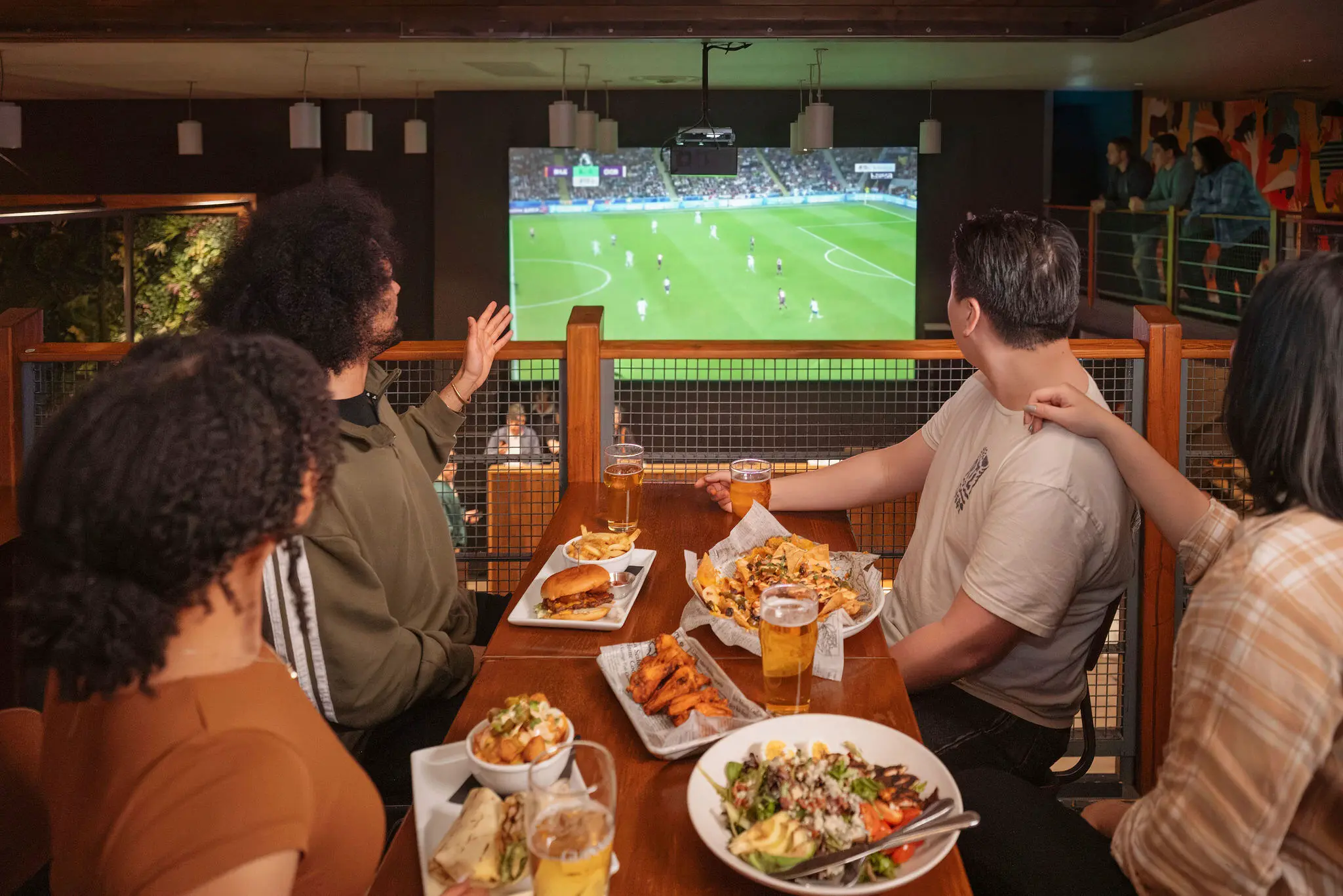 Four people sitting at a table with food and drinks watching a soccer game on a large screen in a restaurant or bar.