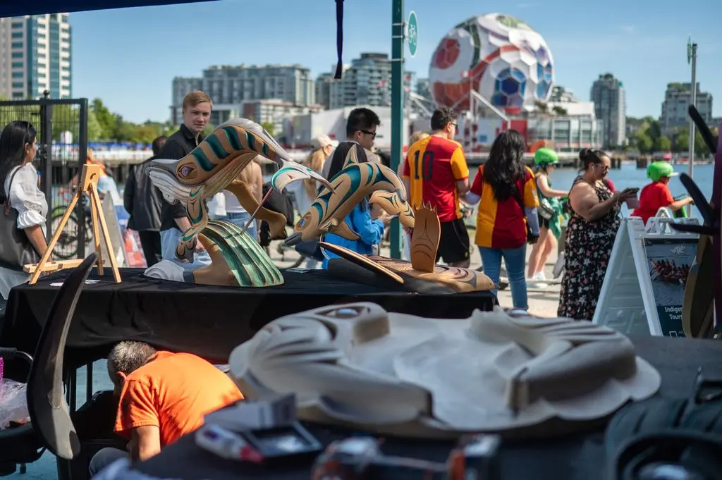 Outdoor market scene with wooden whale sculptures on a table and people walking by near waterfront buildings.