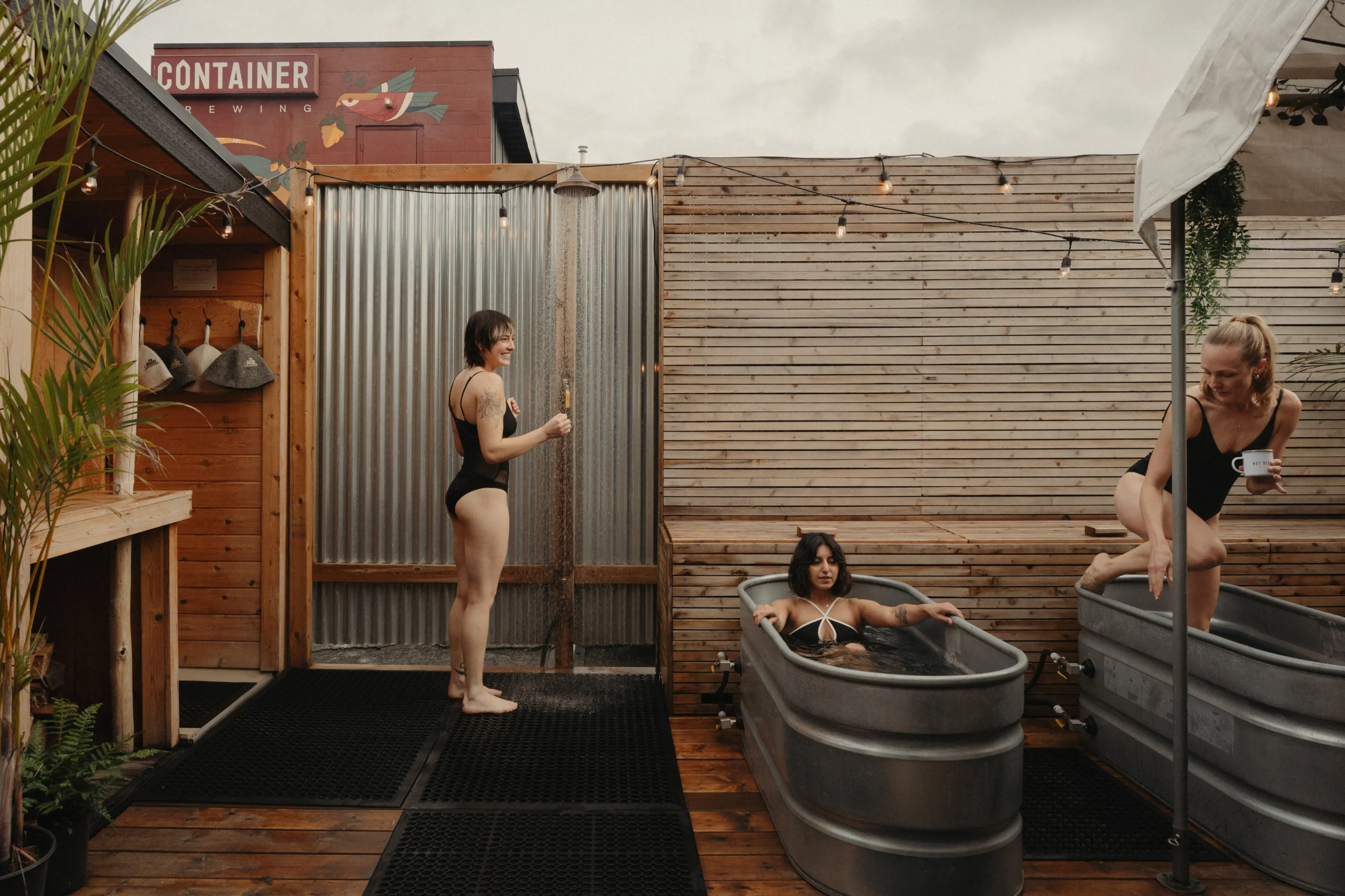 Three women in black swimsuits at an outdoor spa; one showering, one soaking in a metal tub, and one stepping out of another tub holding a cup.