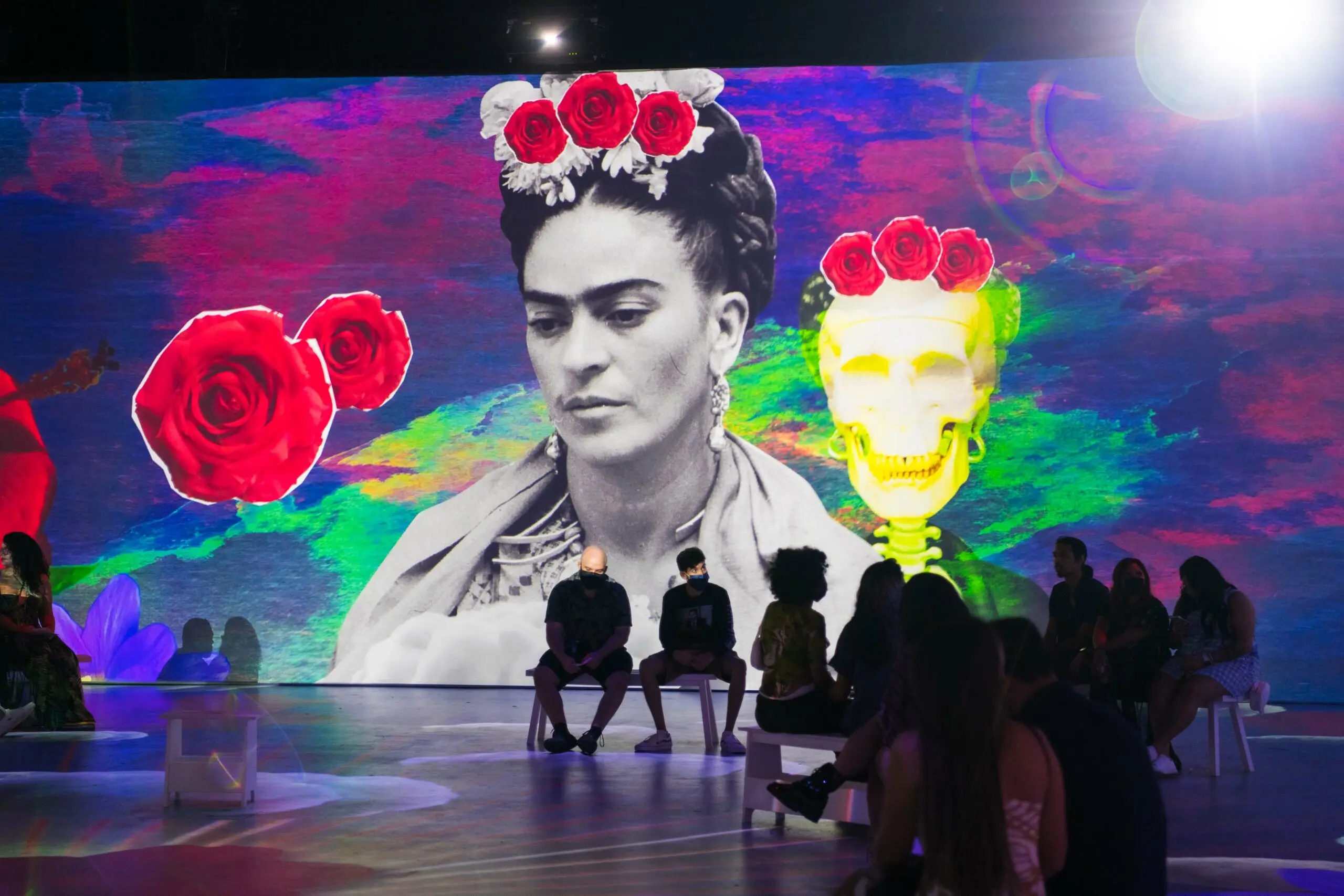 People sitting in front of a large colorful mural featuring a grayscale portrait of a person with flowers on their head and a yellow skeleton with red roses on its head.