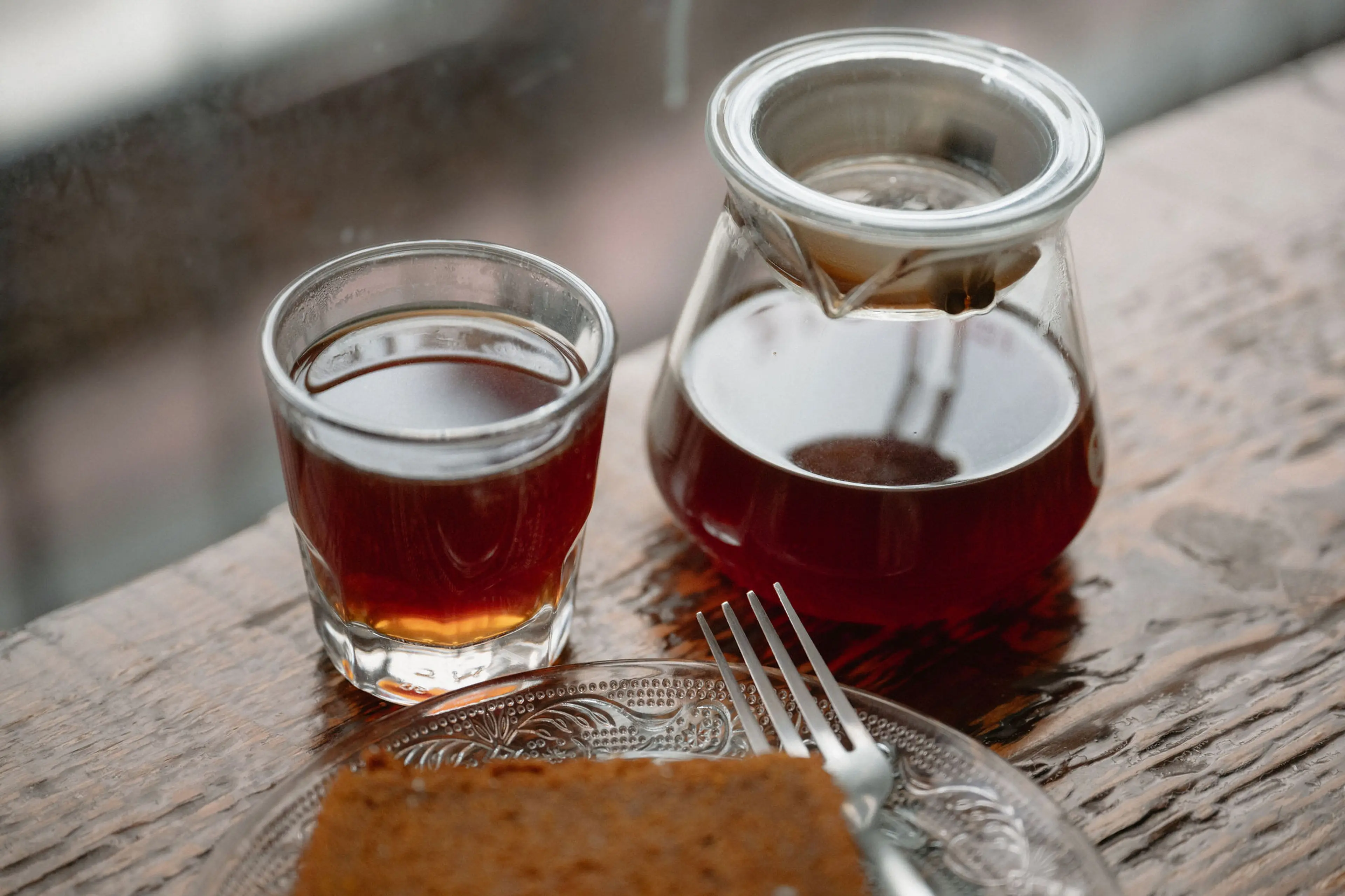 Glass cup and glass pot with dark coffee on a wooden table next to a plate with a fork and a piece of cake.