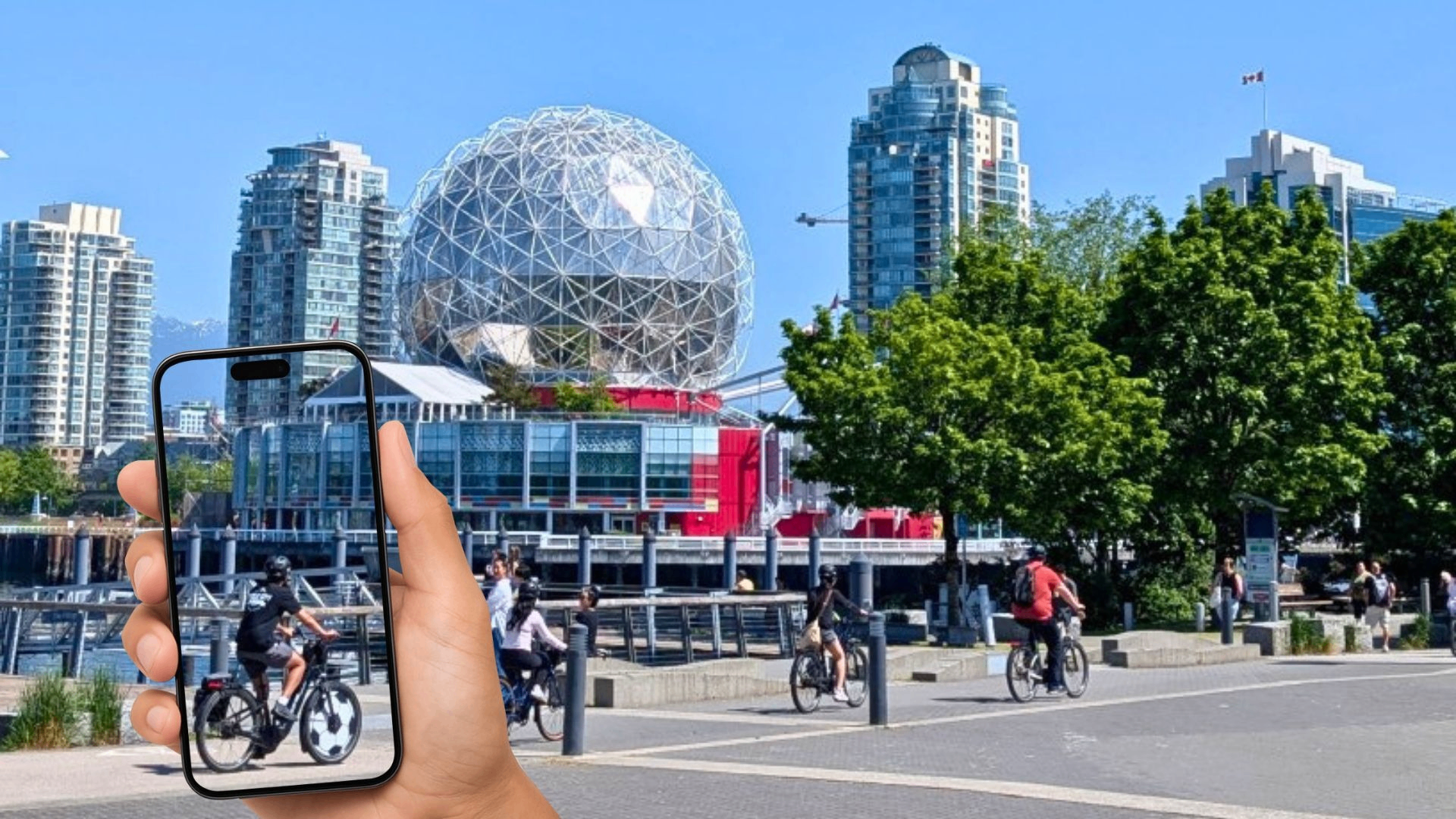 Hand holding a smartphone showing a cyclist, with a waterfront cityscape and a geodesic dome building in the background.