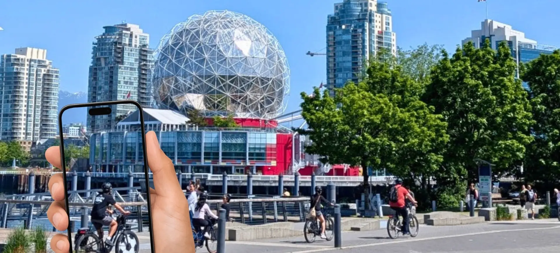 Hand holding a smartphone showing a cyclist, with a waterfront cityscape and a geodesic dome building in the background.