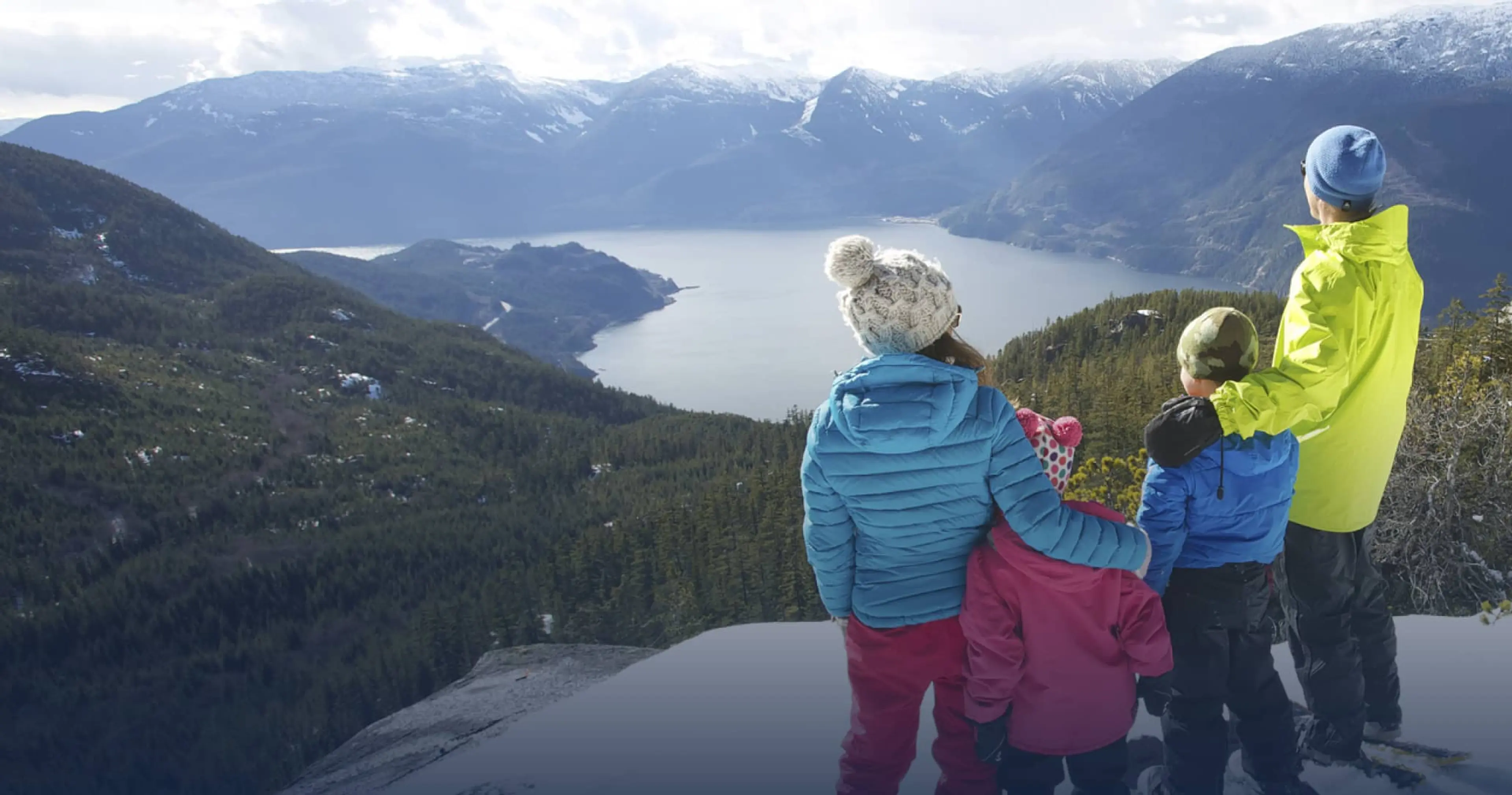 Four people in winter clothing standing on a snowy cliff overlooking a forested valley and a lake with snow-capped mountains in the background.
