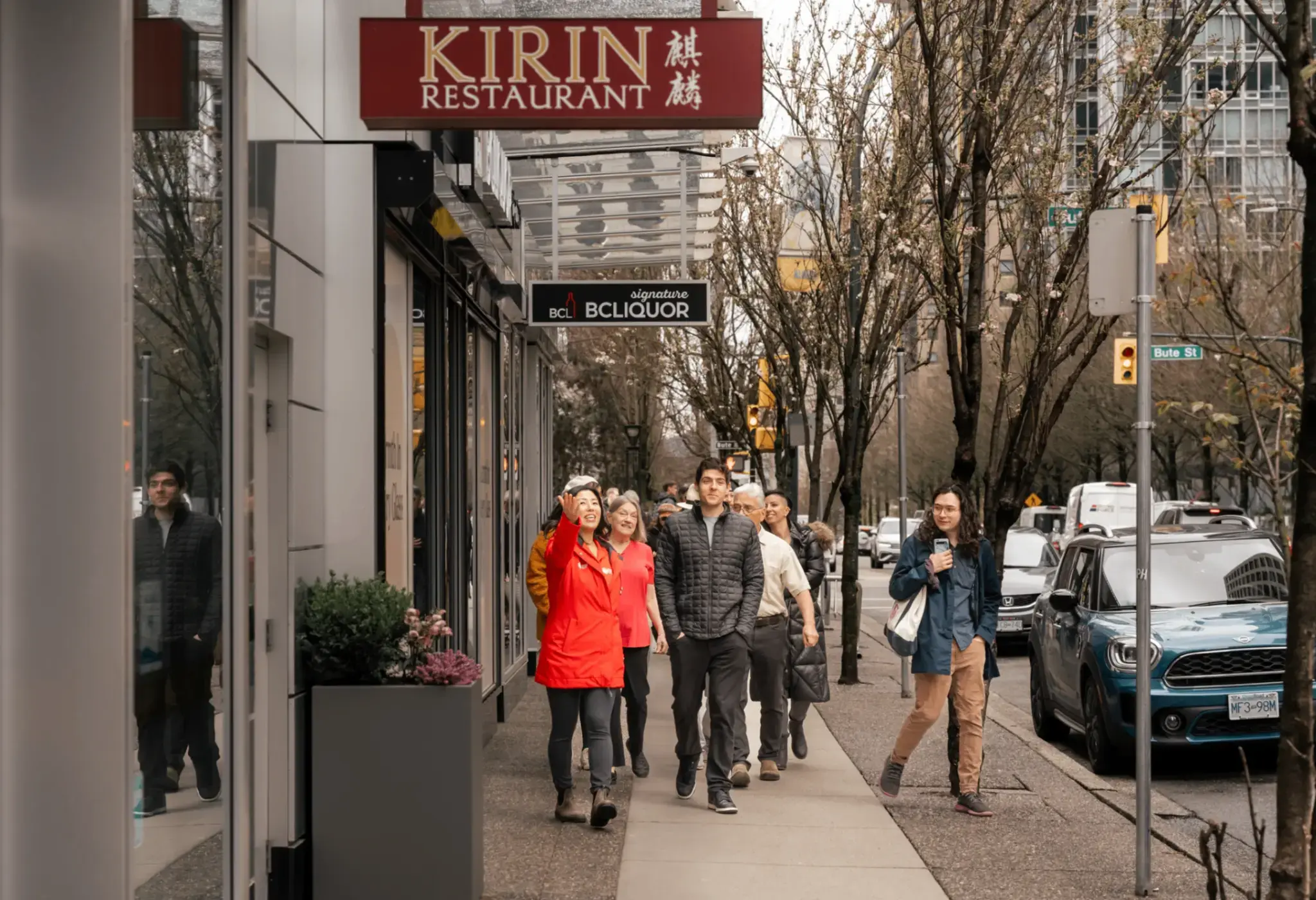 A group of people walking on a city sidewalk past a Kirin Restaurant sign and parked cars on a tree-lined street.