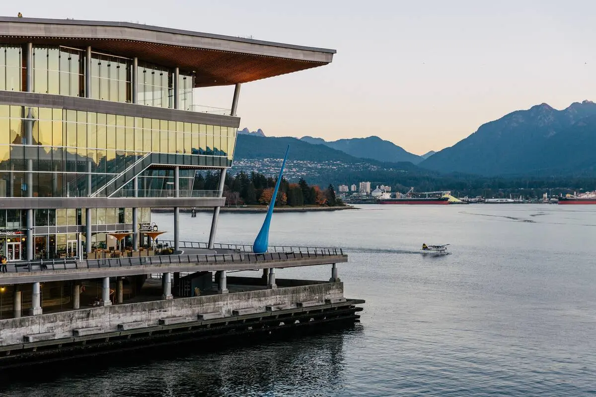 Modern glass building on waterfront with a small boat on water and mountains in the background at sunset.