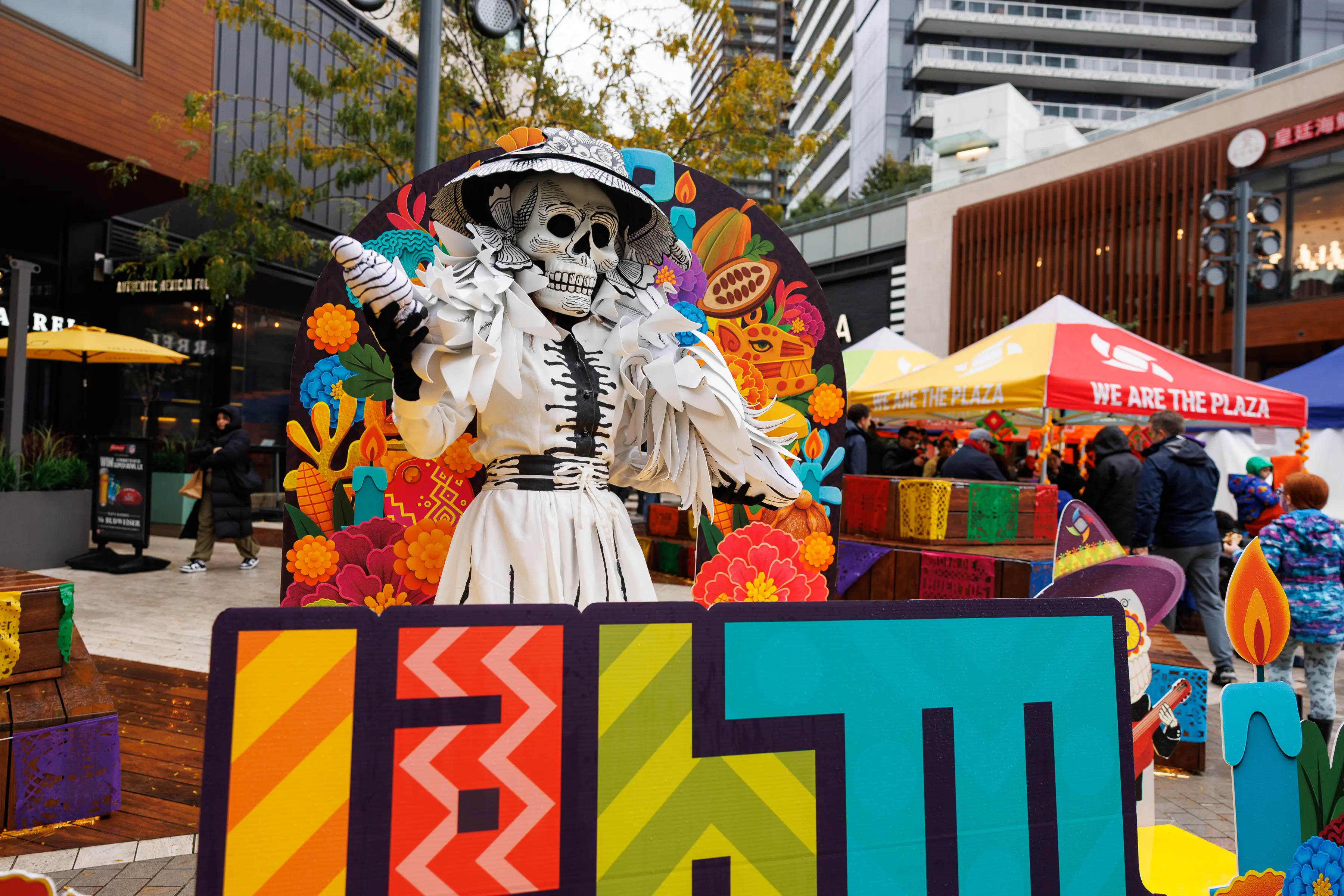 Skeleton figure in white dress and hat with colorful floral and candle decorations behind, at an outdoor plaza event.