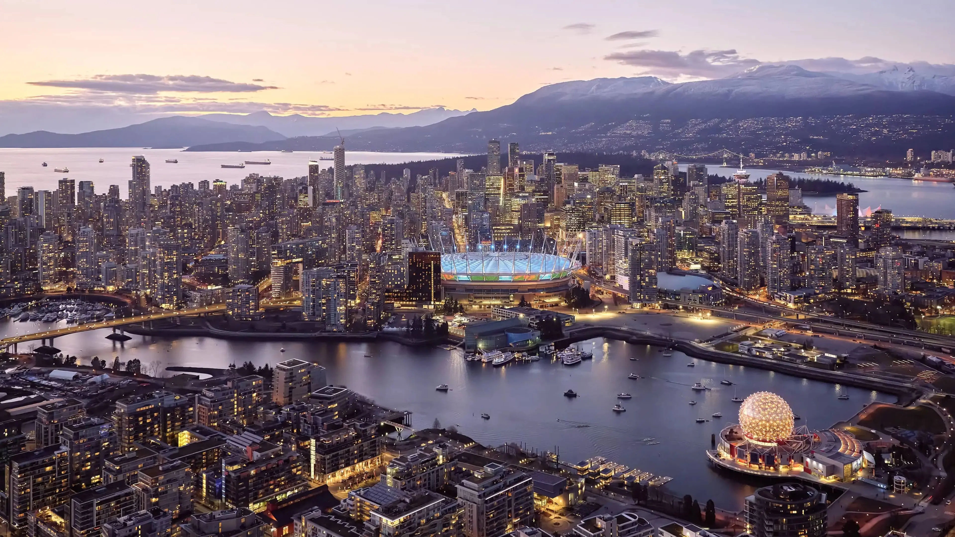 Dusk aerial view of Vancouver with BC Place glowing softly in the distance as the city transitions into night.