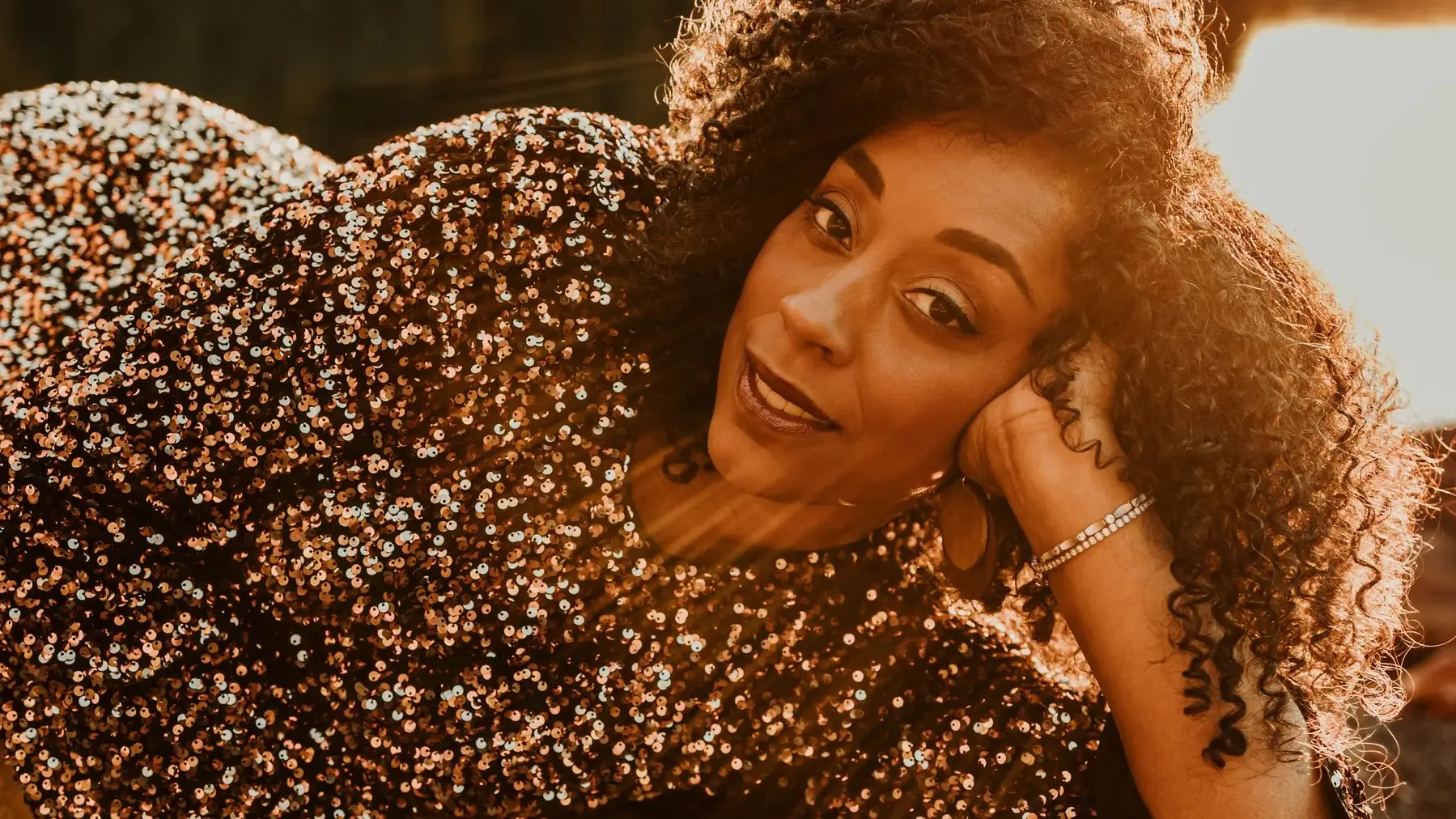 Person with curly hair wearing a sequined top, resting head on hand with sunlight shining from behind.