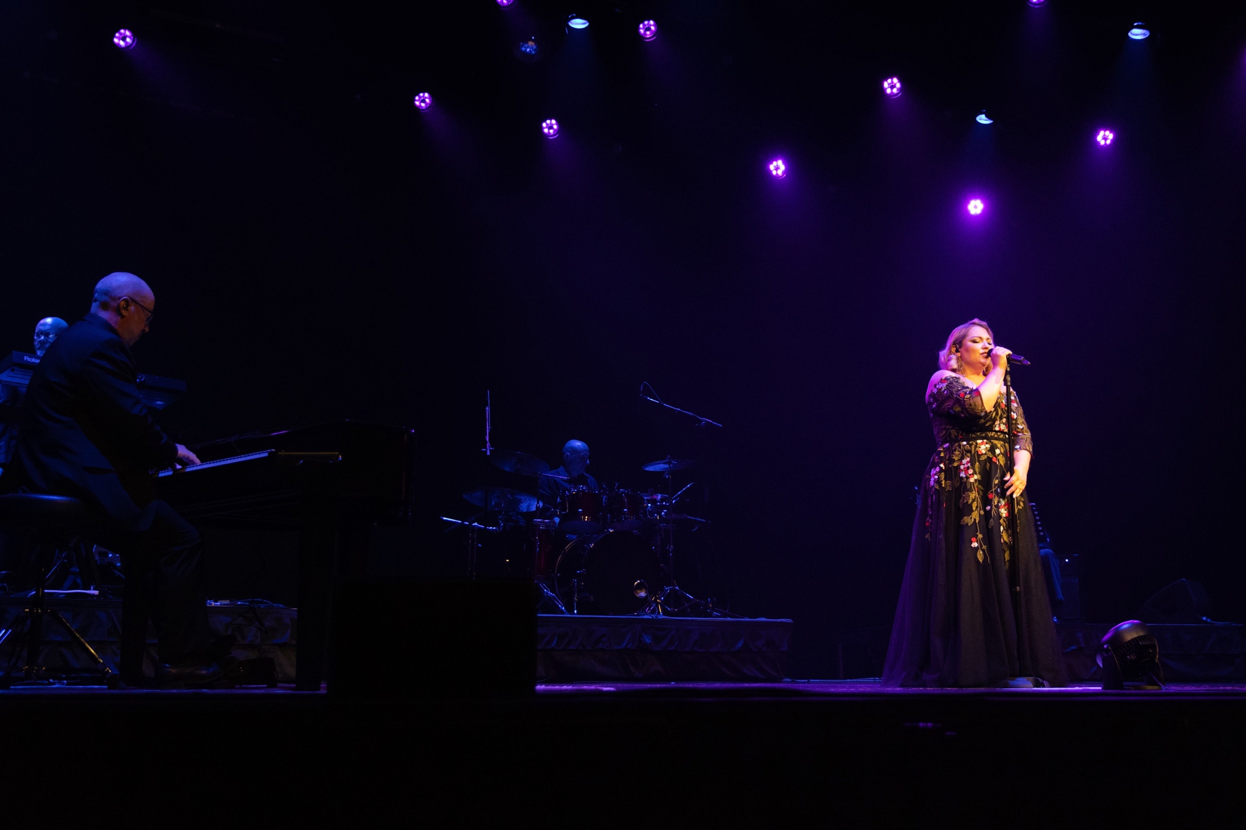 A person in a floral dress singing on stage with purple spotlights, accompanied by musicians playing piano and drums.