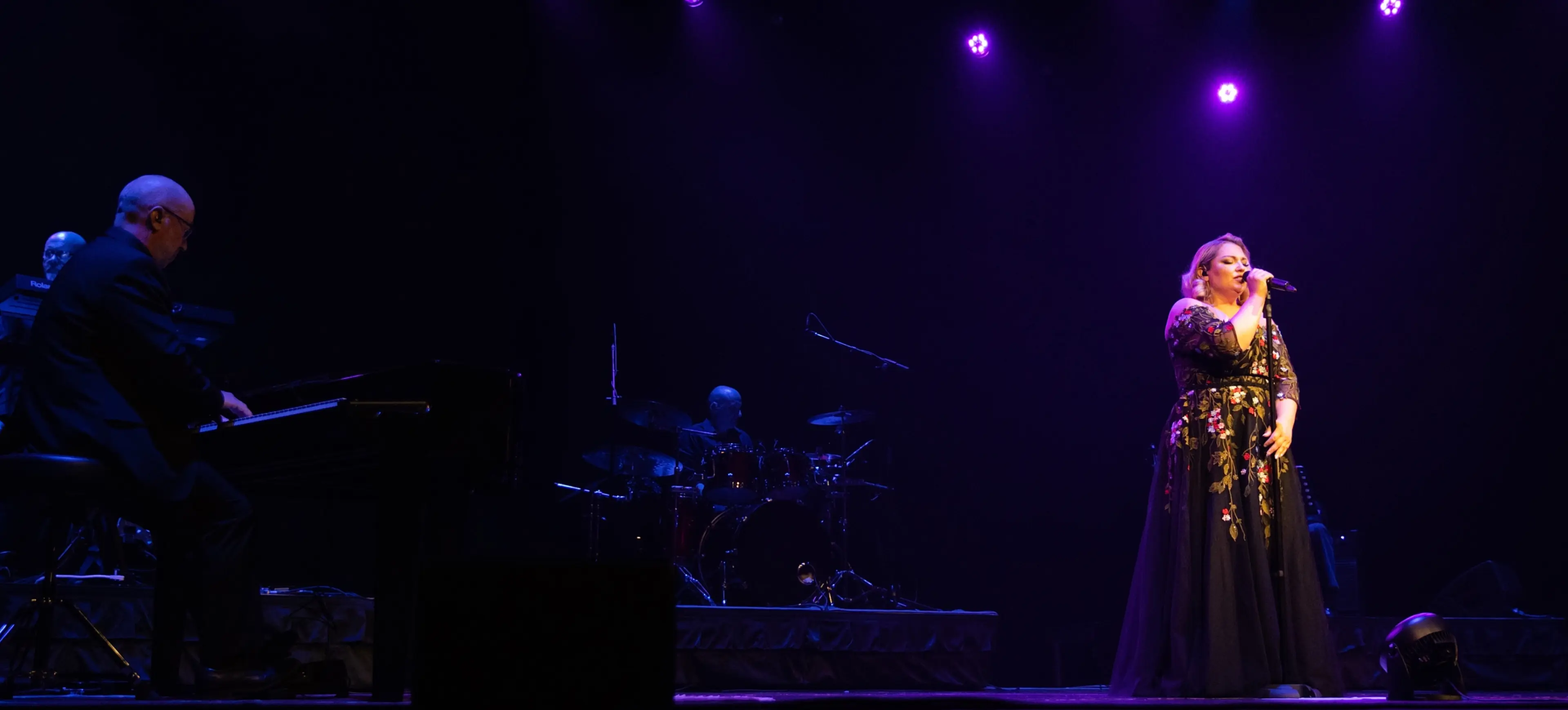 A person in a floral dress singing on stage with purple spotlights, accompanied by musicians playing piano and drums.