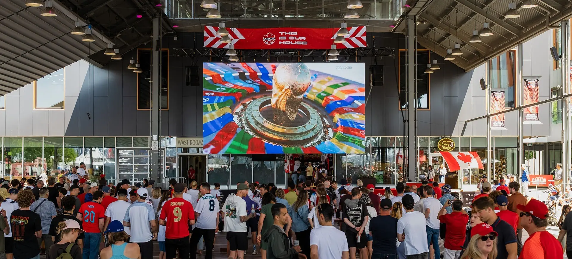 Crowd of people watching a large screen showing a World Cup trophy inside a covered outdoor area with Canadian flags and a sign reading "This is our house".
