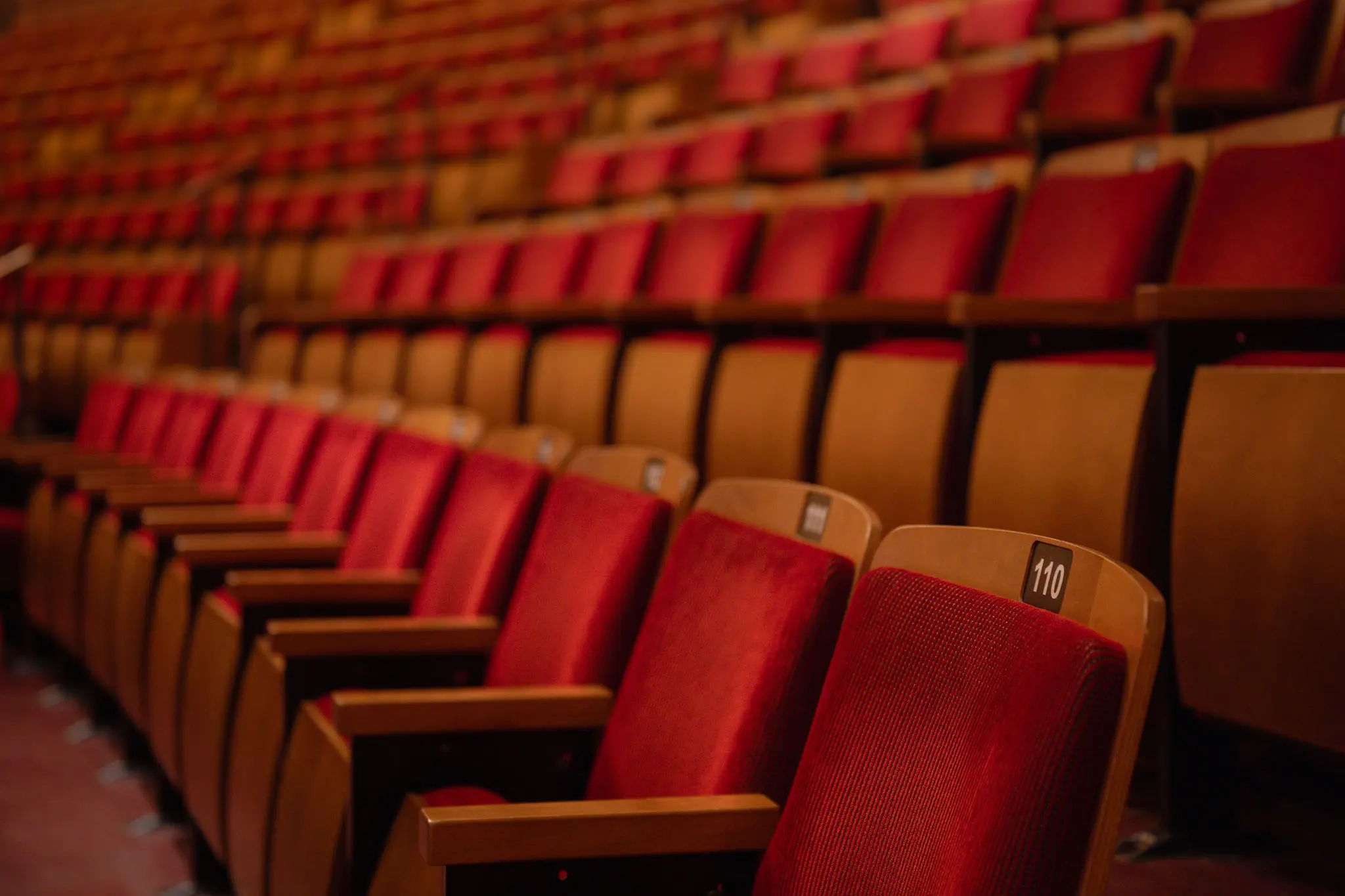Rows of empty theater seats with red cushions and wooden armrests, seat number 110 visible.