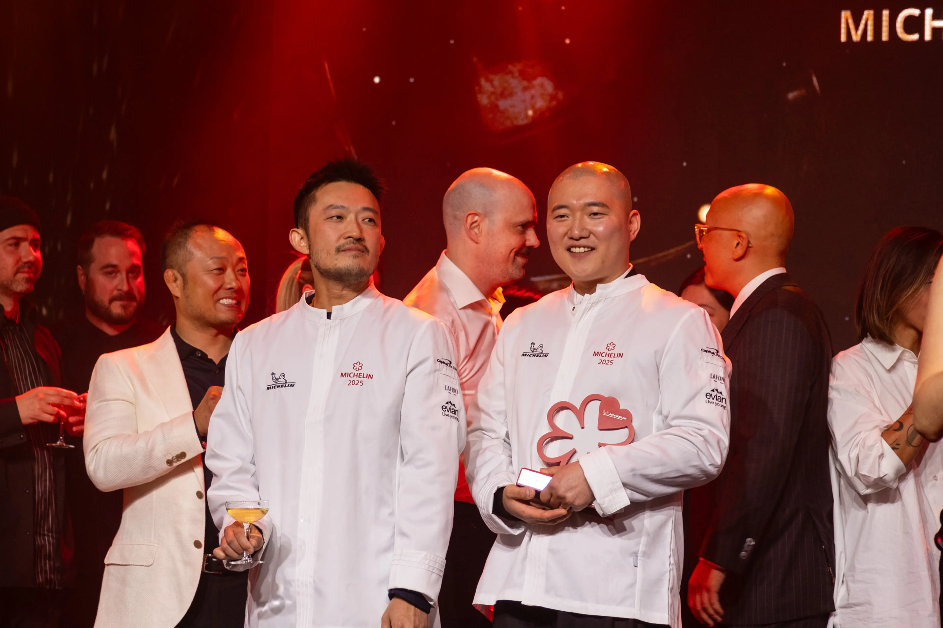 Two chefs in white Michelin 2025 jackets holding a Michelin star award, surrounded by people in formal attire at an event.