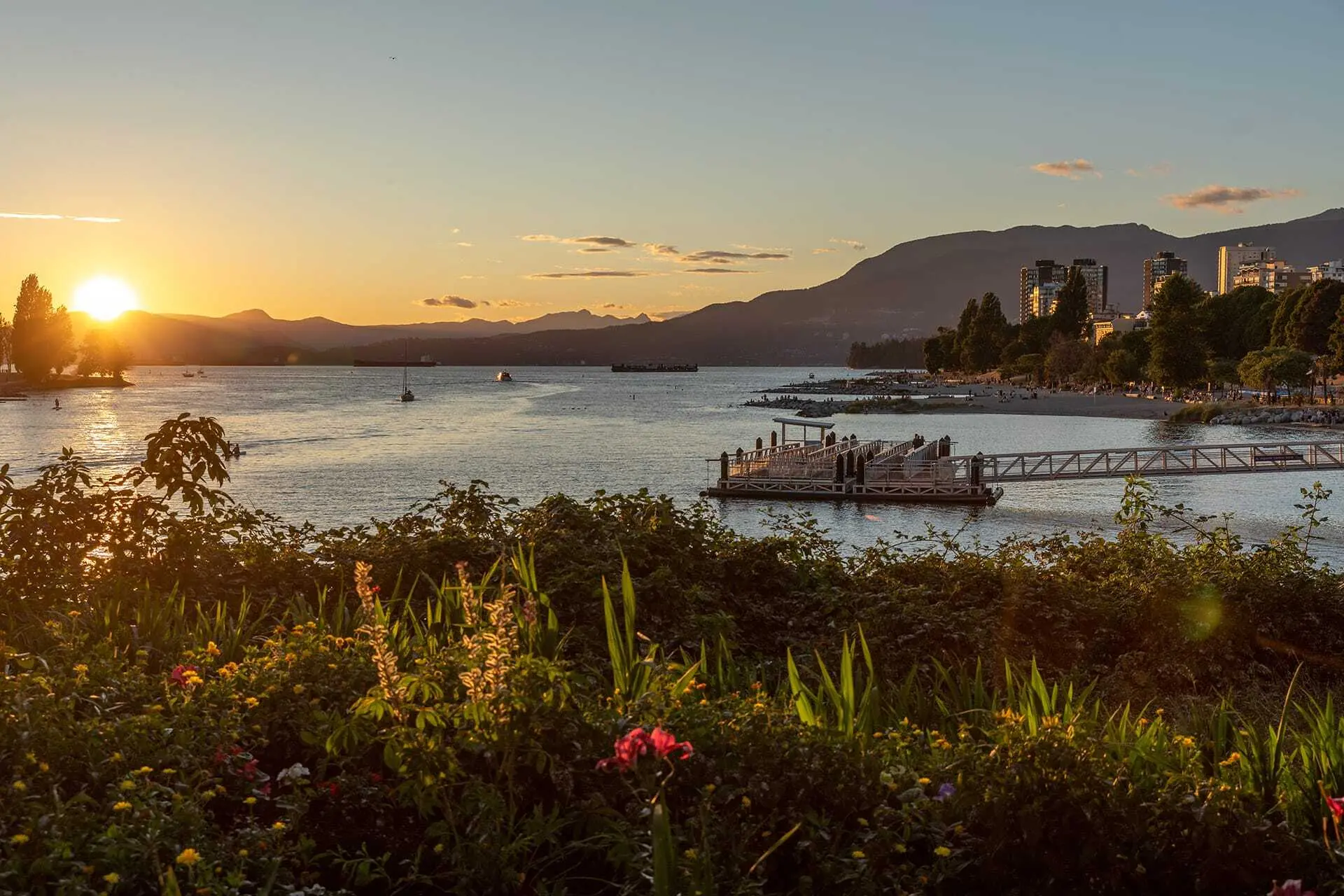 Sunset over a calm bay with a dock, boats on the water, city buildings, and mountains in the background.