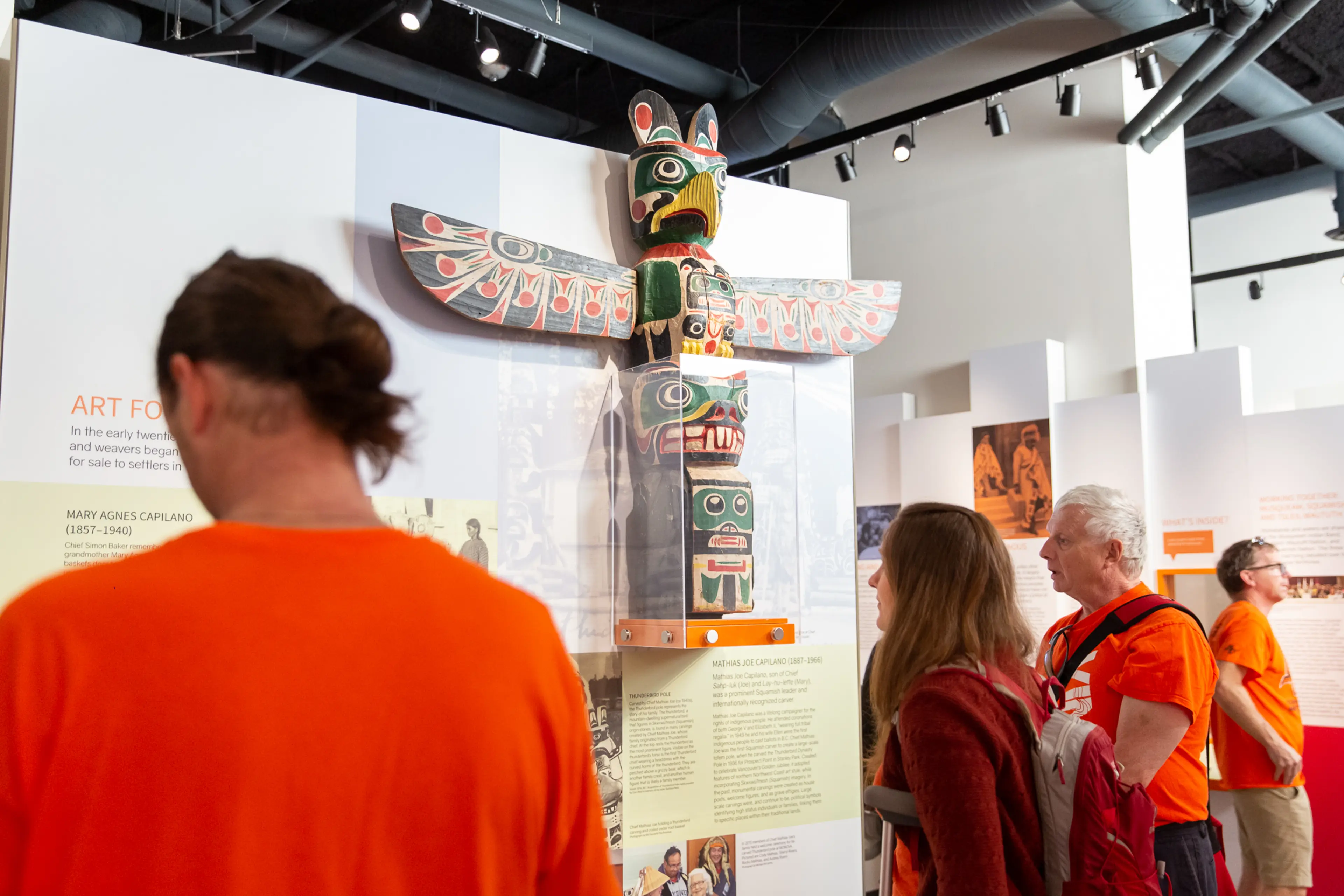 Four people observing a colorful totem pole displayed in a museum exhibit with informational panels.