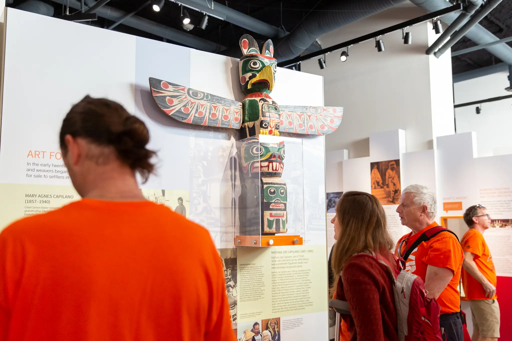 Four people observing a colorful totem pole displayed in a museum exhibit with informational panels.