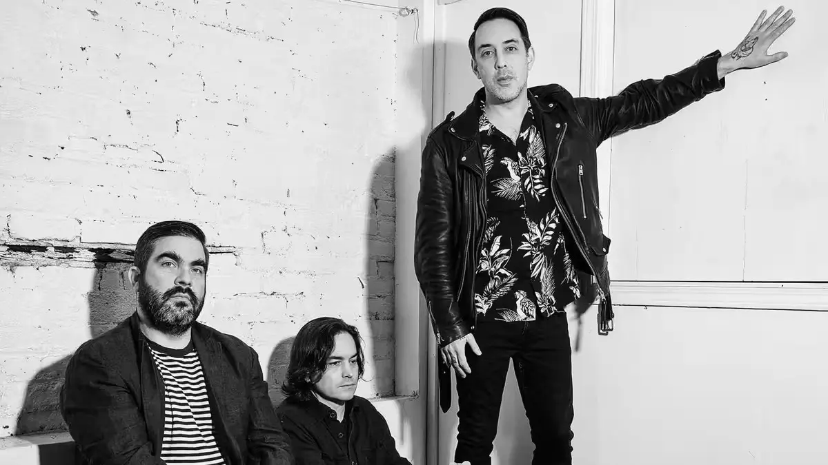 Three people posing against a white brick wall, one standing with arm extended, two sitting, all wearing dark clothing.