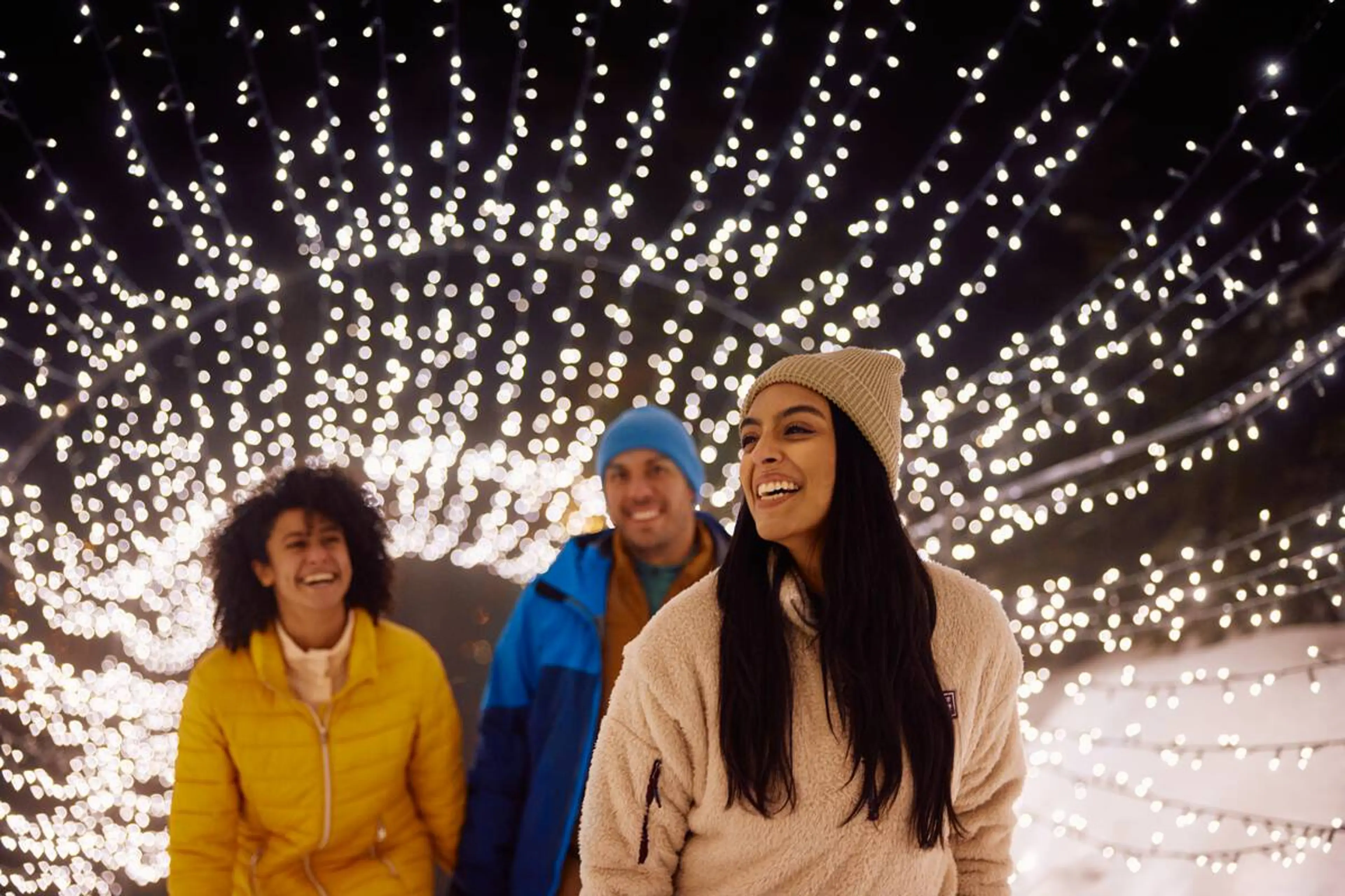 Three people smile as they walk through a tunnel of lights in the snow at Grouse Mountain.