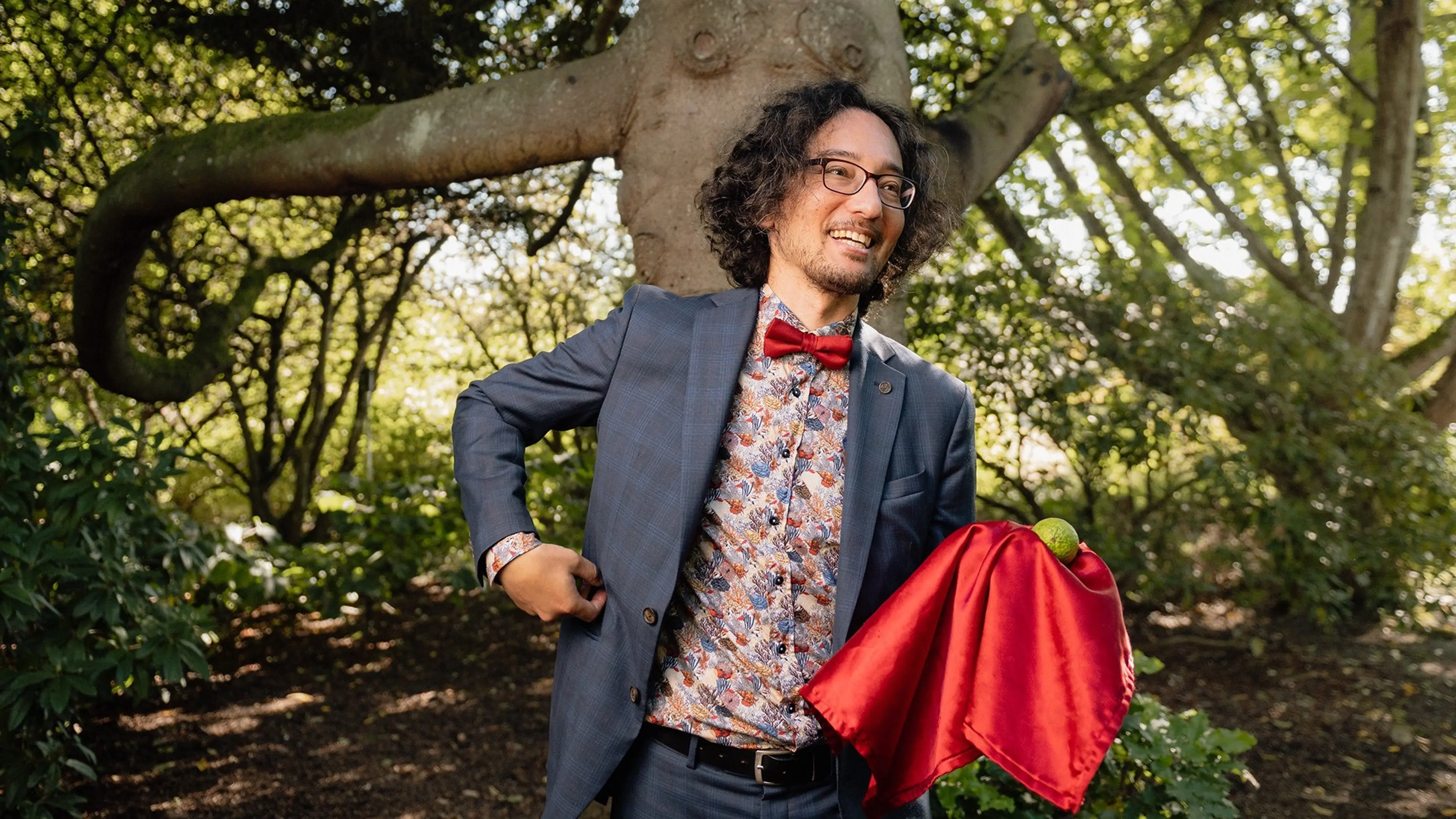 Person with curly hair and glasses wearing a blue suit and red bow tie, holding a red cloth with a green ball outdoors.