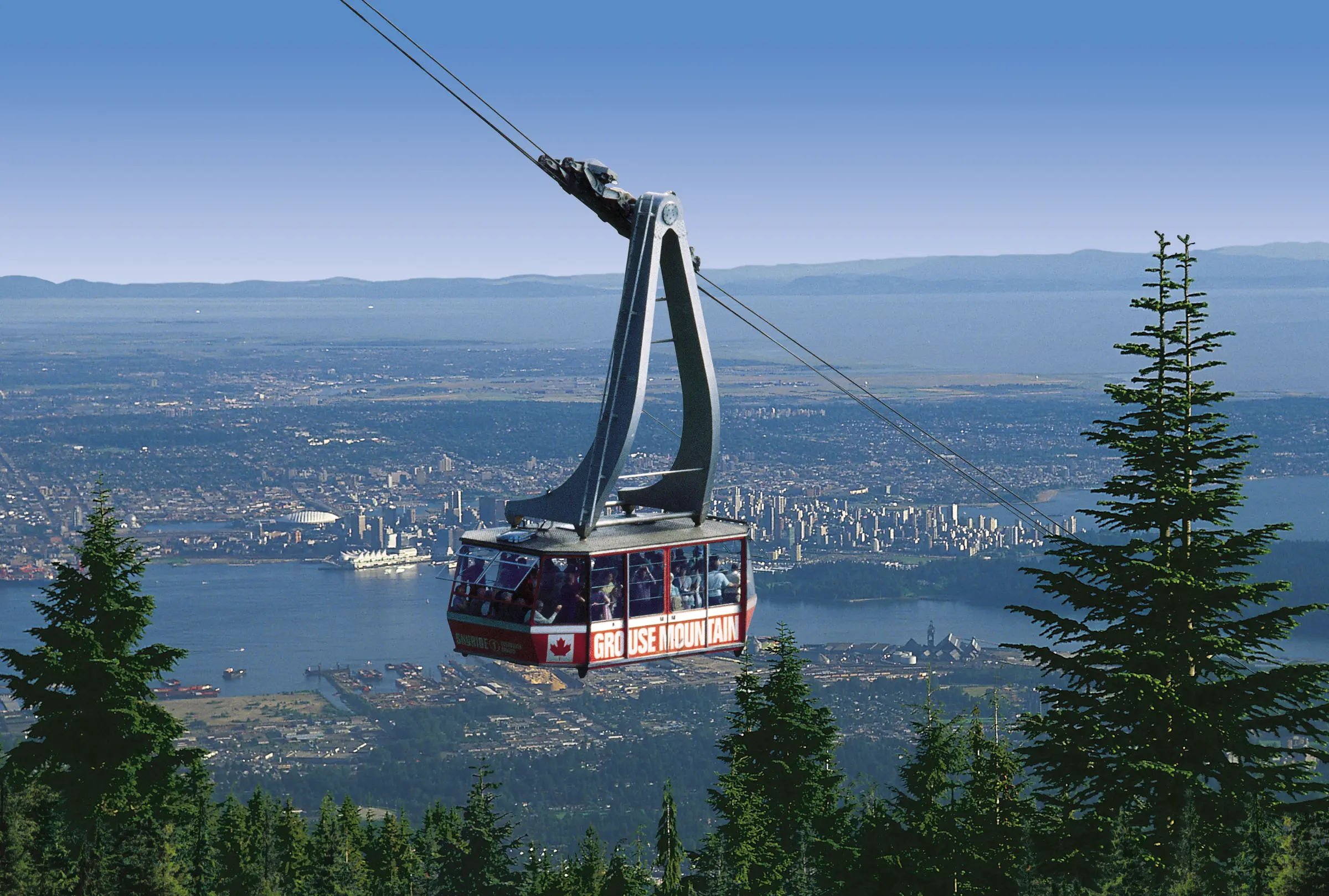 Gondola labeled Grouse Mountain carrying passengers over forest with city and water in the background