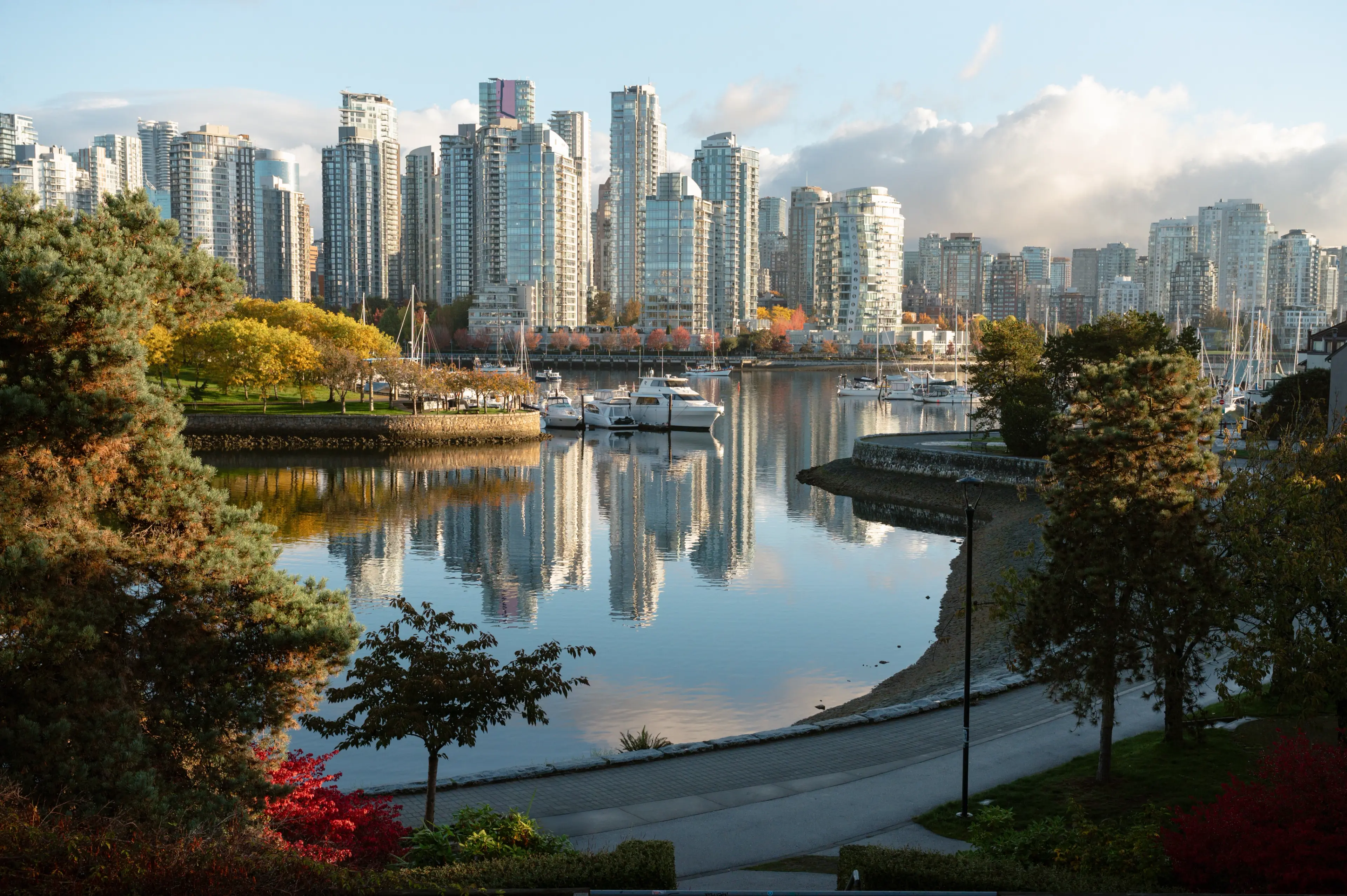 City skyline with tall glass buildings reflecting on calm water, surrounded by trees and a walkway.