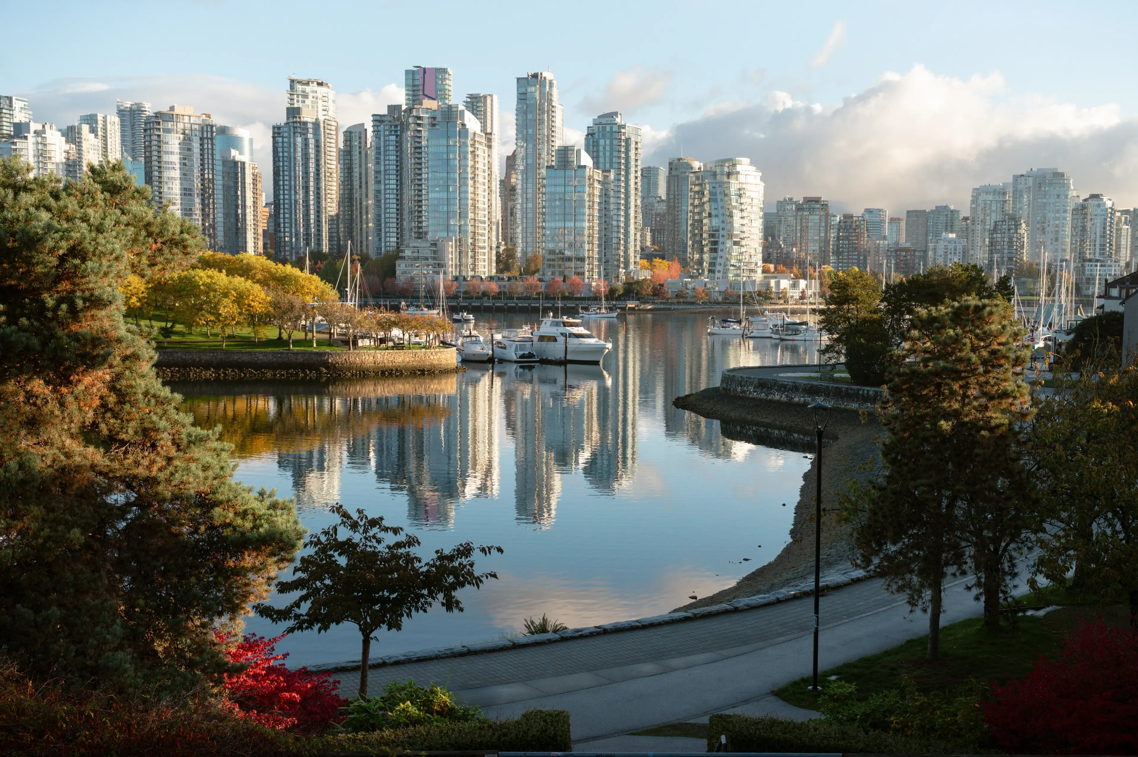 City skyline with tall glass buildings reflecting on calm water, surrounded by trees and a walkway.