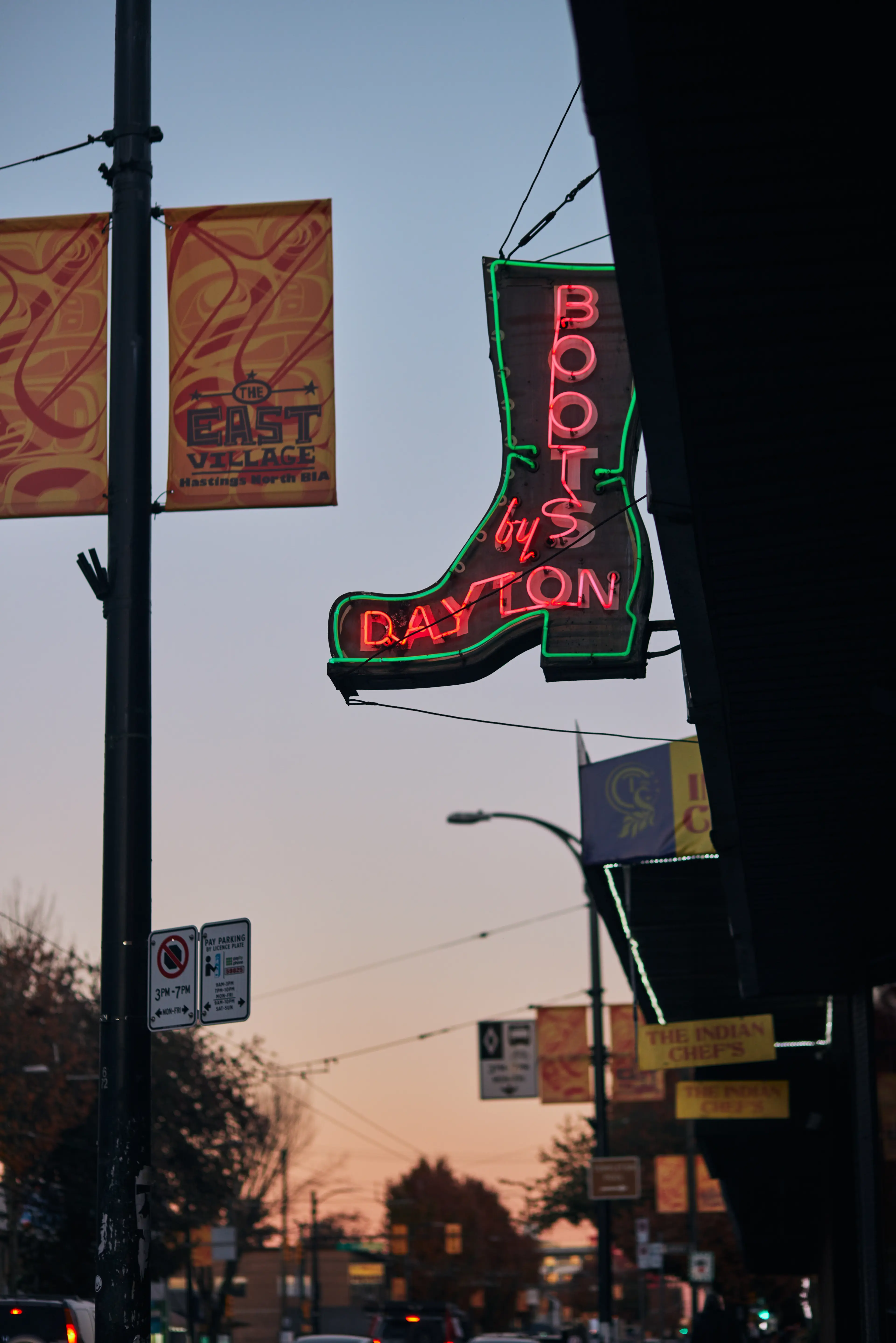 Neon sign shaped like a boot reads 'Boots by Dayton' on a city street at dusk.
