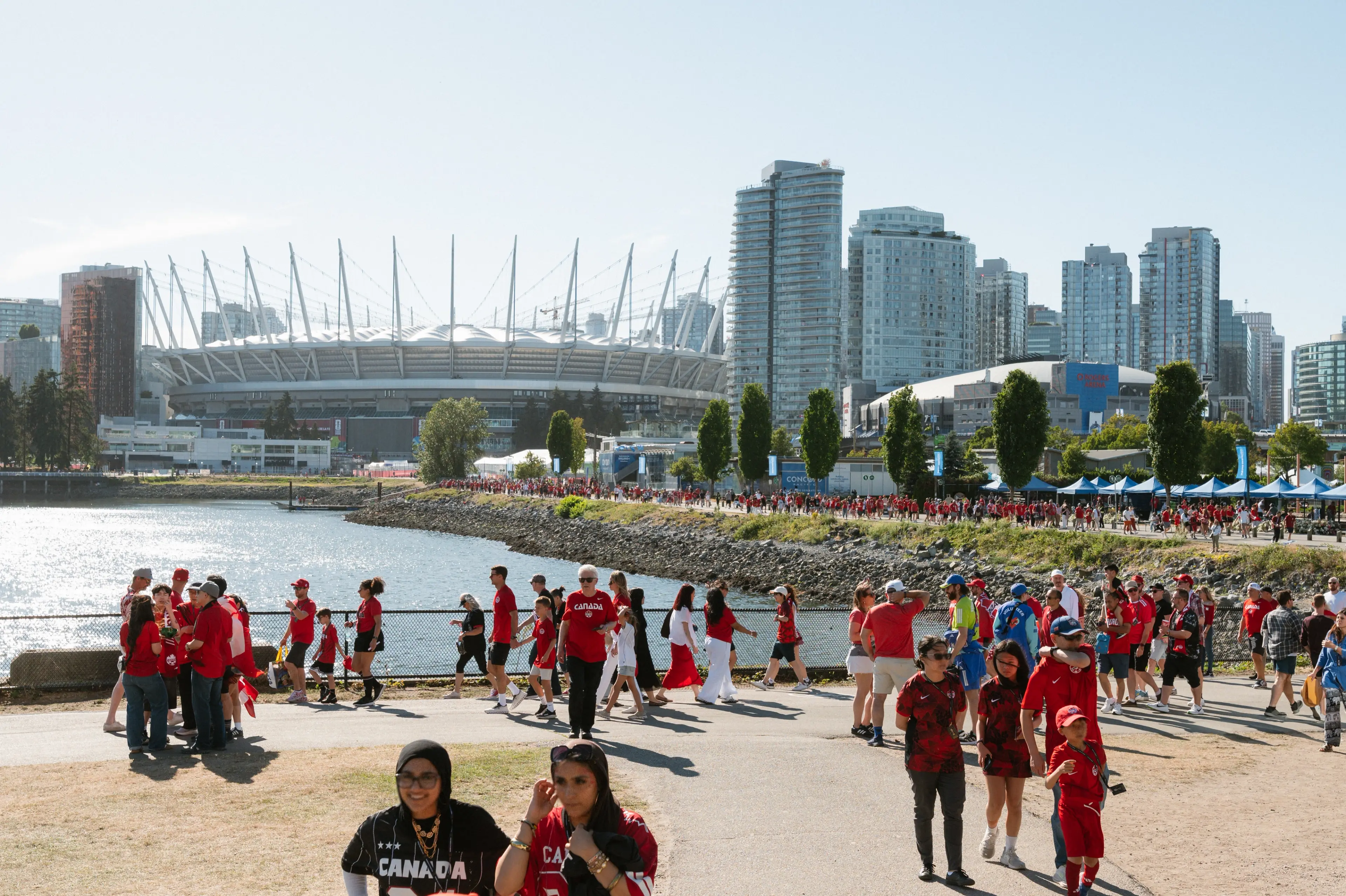 Crowd of people wearing red near waterfront with stadium and city skyline in the background on a sunny day.