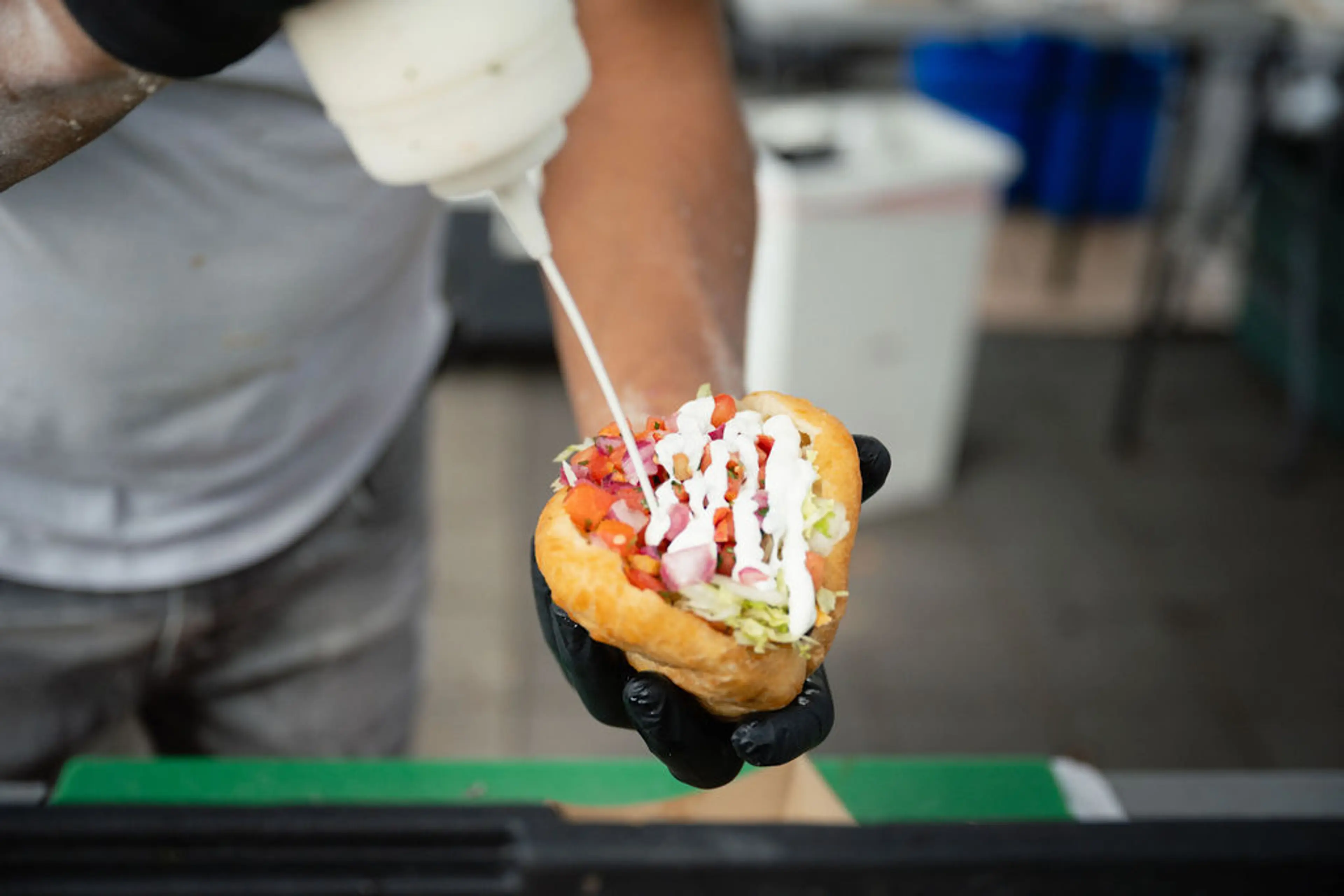 Person wearing black gloves drizzling white sauce on a sandwich filled with lettuce, tomatoes, and other vegetables.