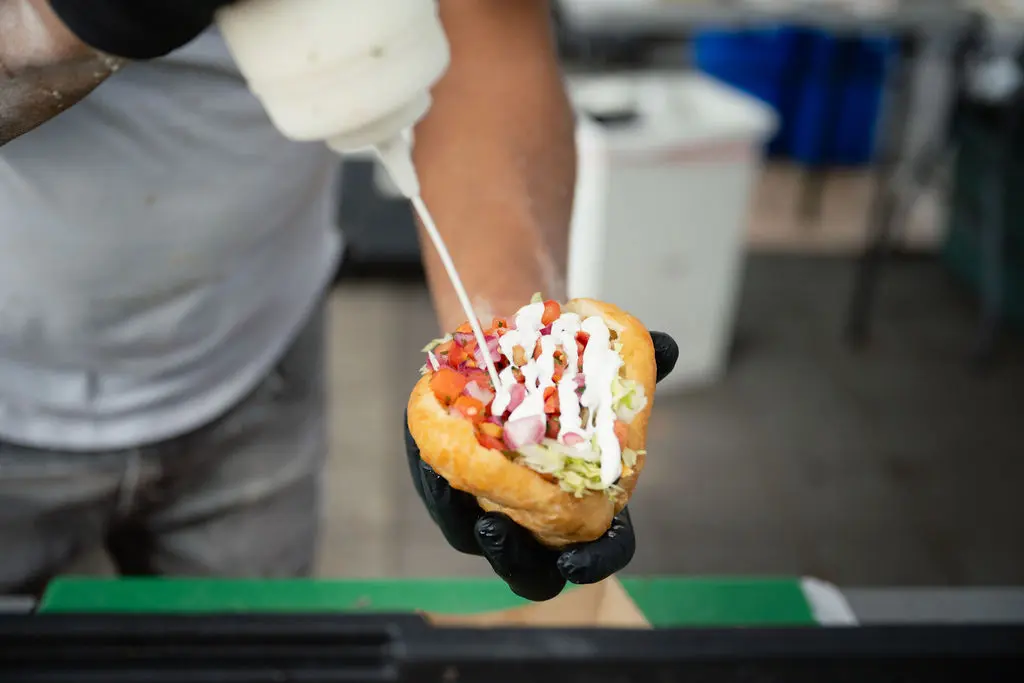 Person wearing black gloves drizzling white sauce on a sandwich filled with lettuce, tomatoes, and other vegetables.