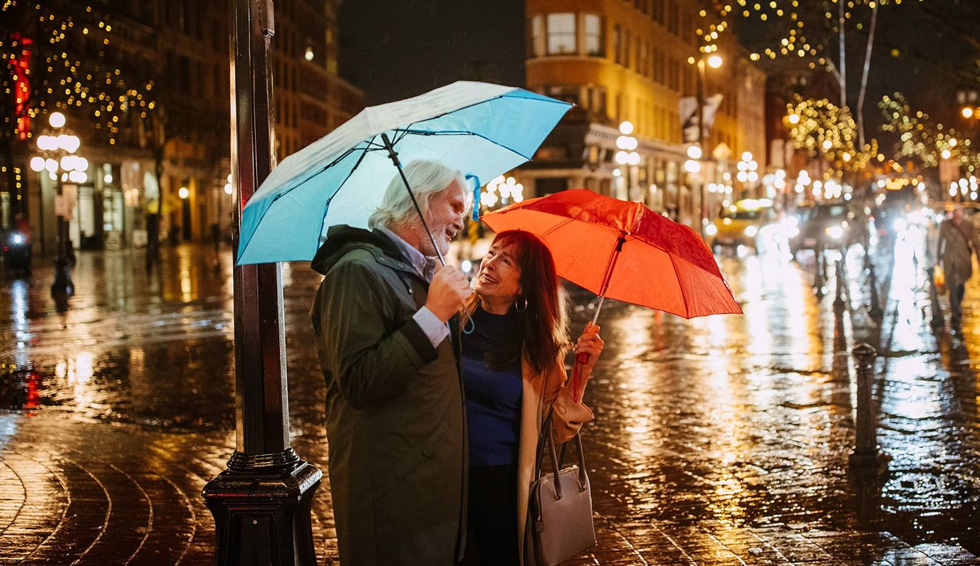 Two people standing close under umbrellas on a wet city street at night with lights reflecting on the pavement.