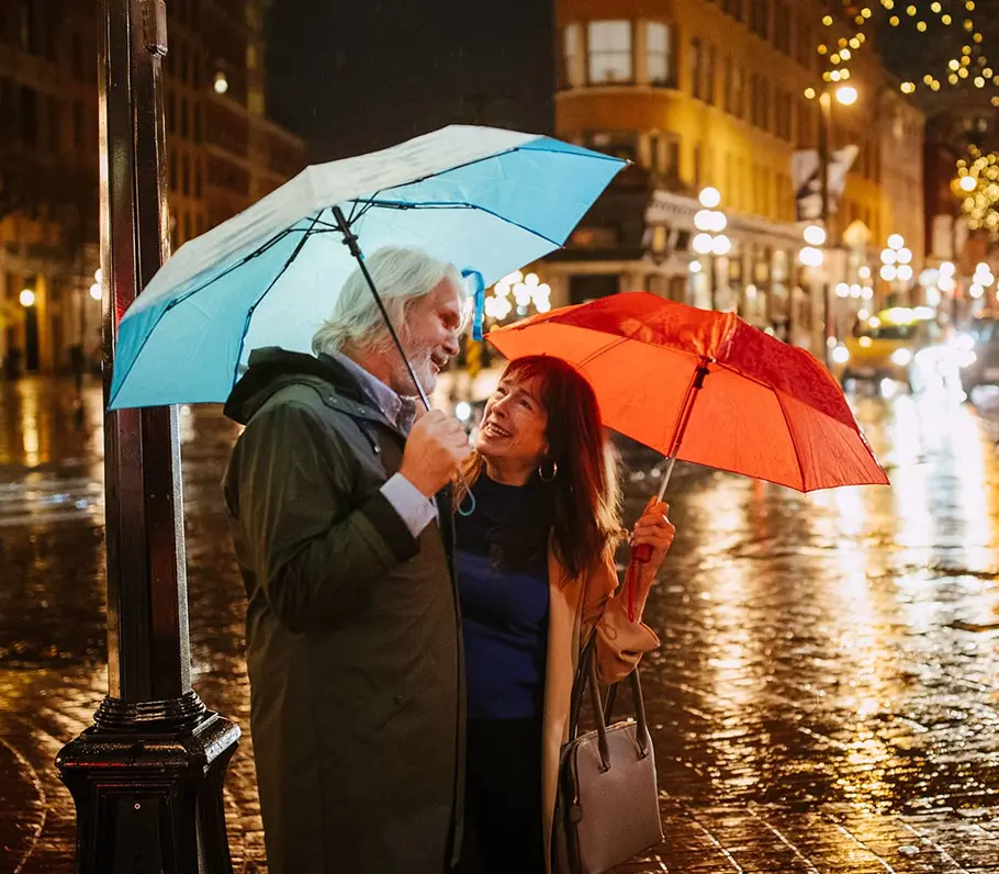 Two people standing close under umbrellas on a wet city street at night with lights reflecting on the pavement.