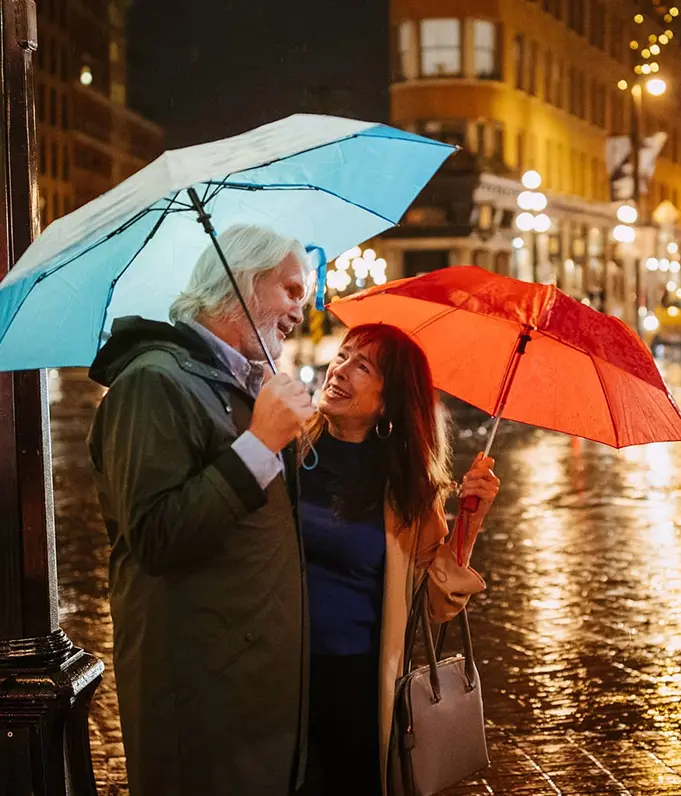 Two people standing close under umbrellas on a wet city street at night with lights reflecting on the pavement.