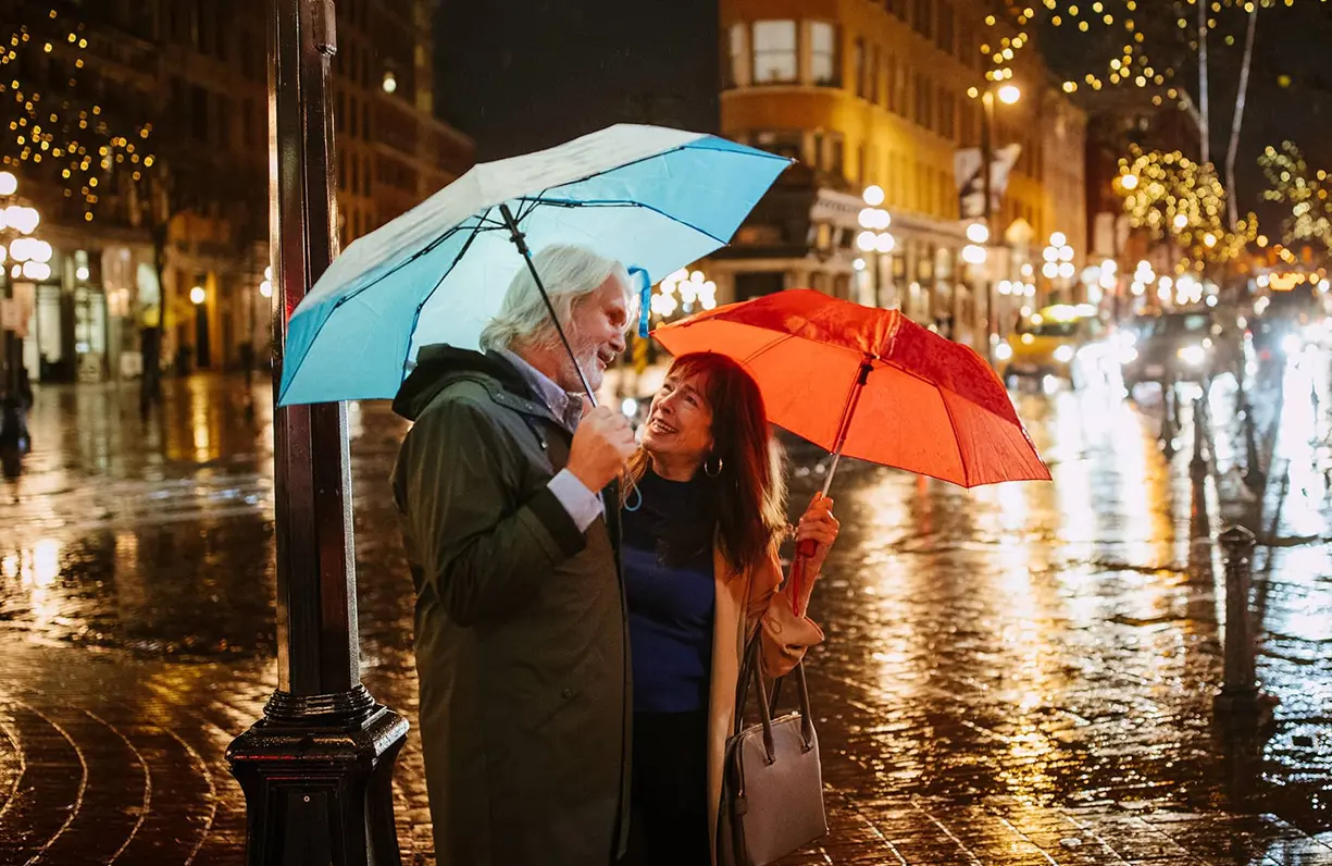 Two people standing close under umbrellas on a wet city street at night with lights reflecting on the pavement.