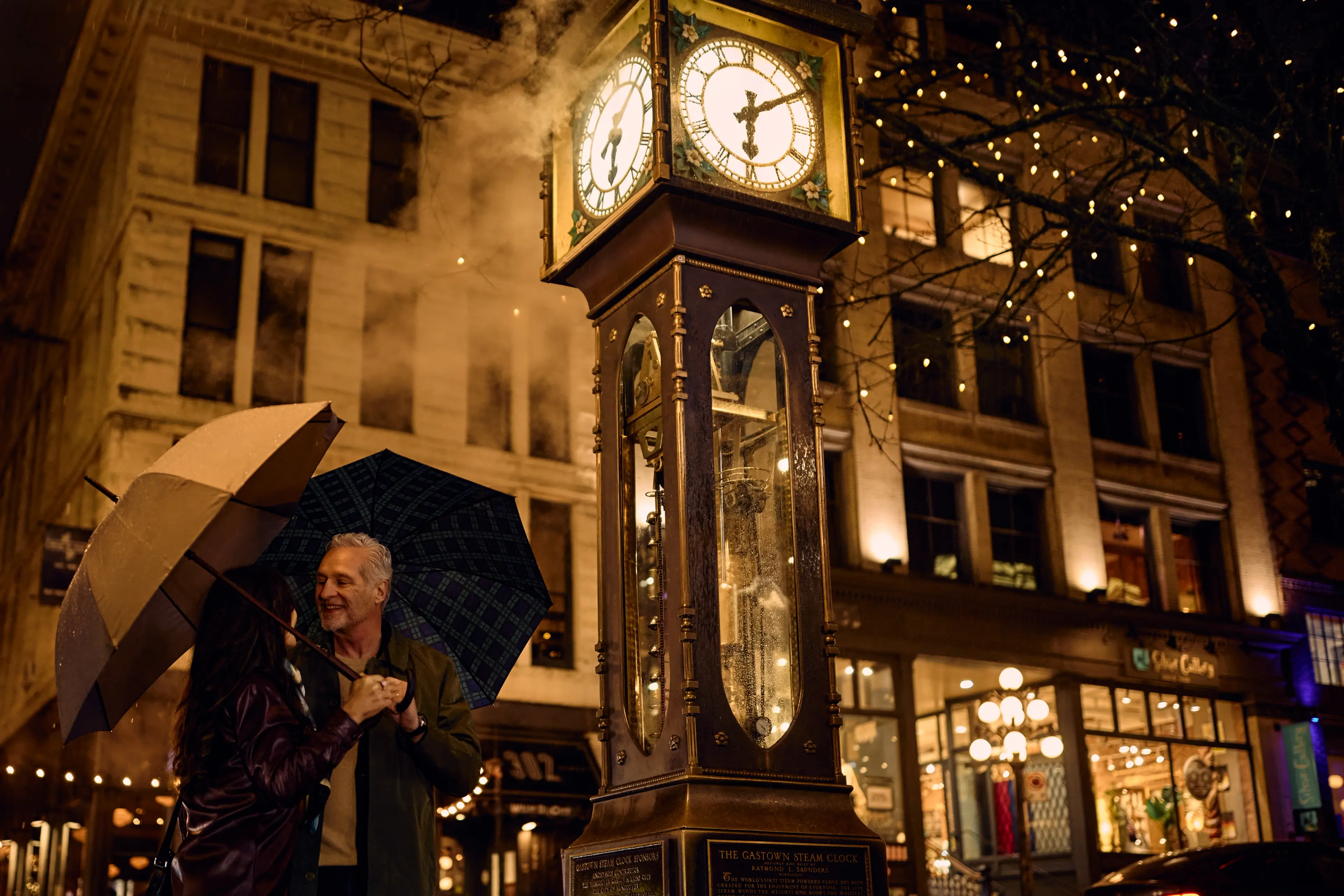 A romantic scene of a couple standing in front of the Gastown Steam Clock, holding umbrellas beneath twinkling lights.