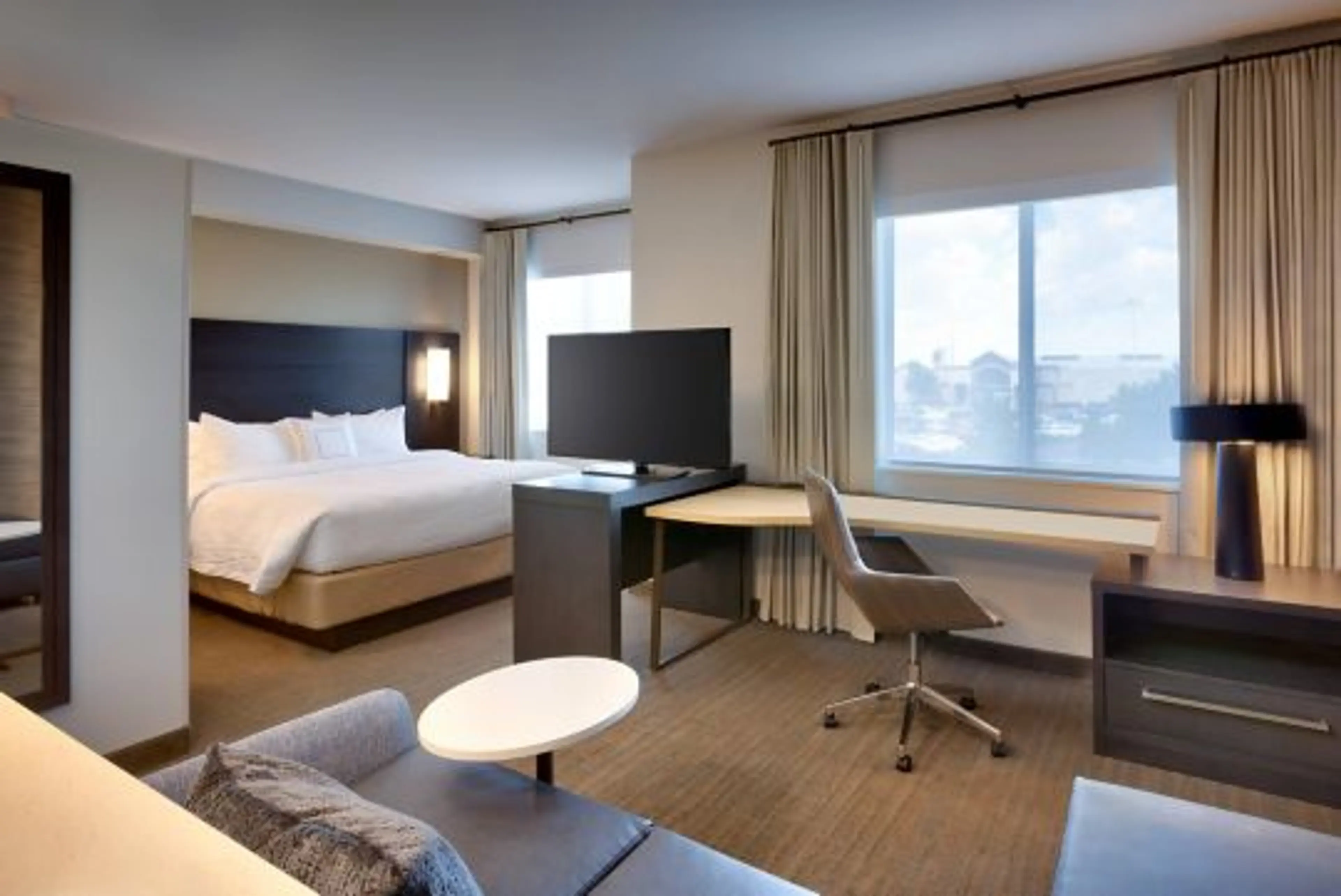 Hotel room with a large bed, TV on a stand, desk with chair, window with curtains, and a small round table next to a sofa.
