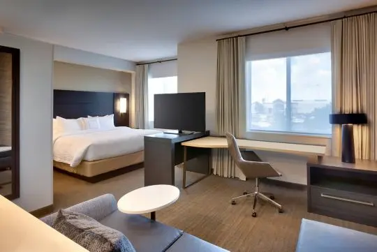 Hotel room with a large bed, TV on a stand, desk with chair, window with curtains, and a small round table next to a sofa.