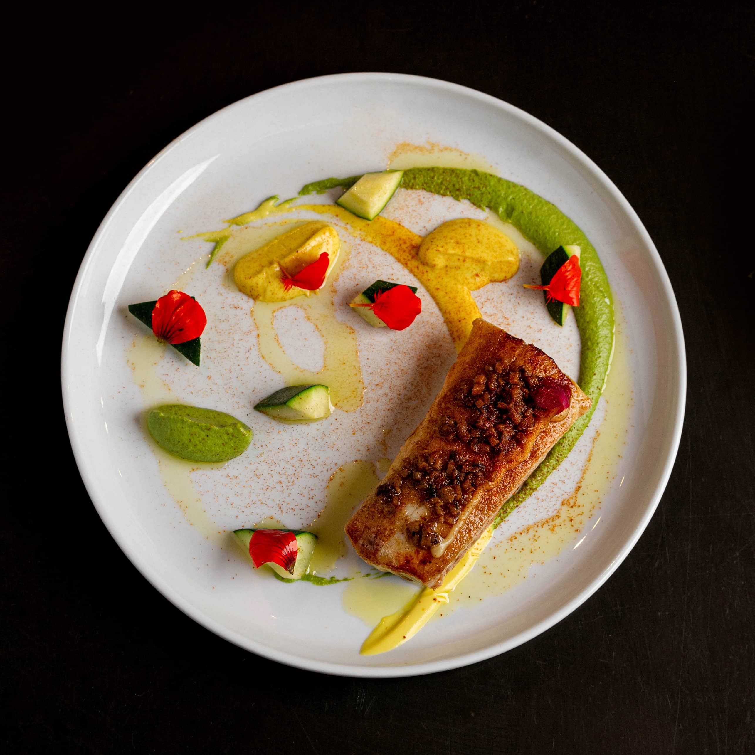 Grilled fish fillet on a white plate with green and yellow sauces, cucumber pieces, and red edible flowers.