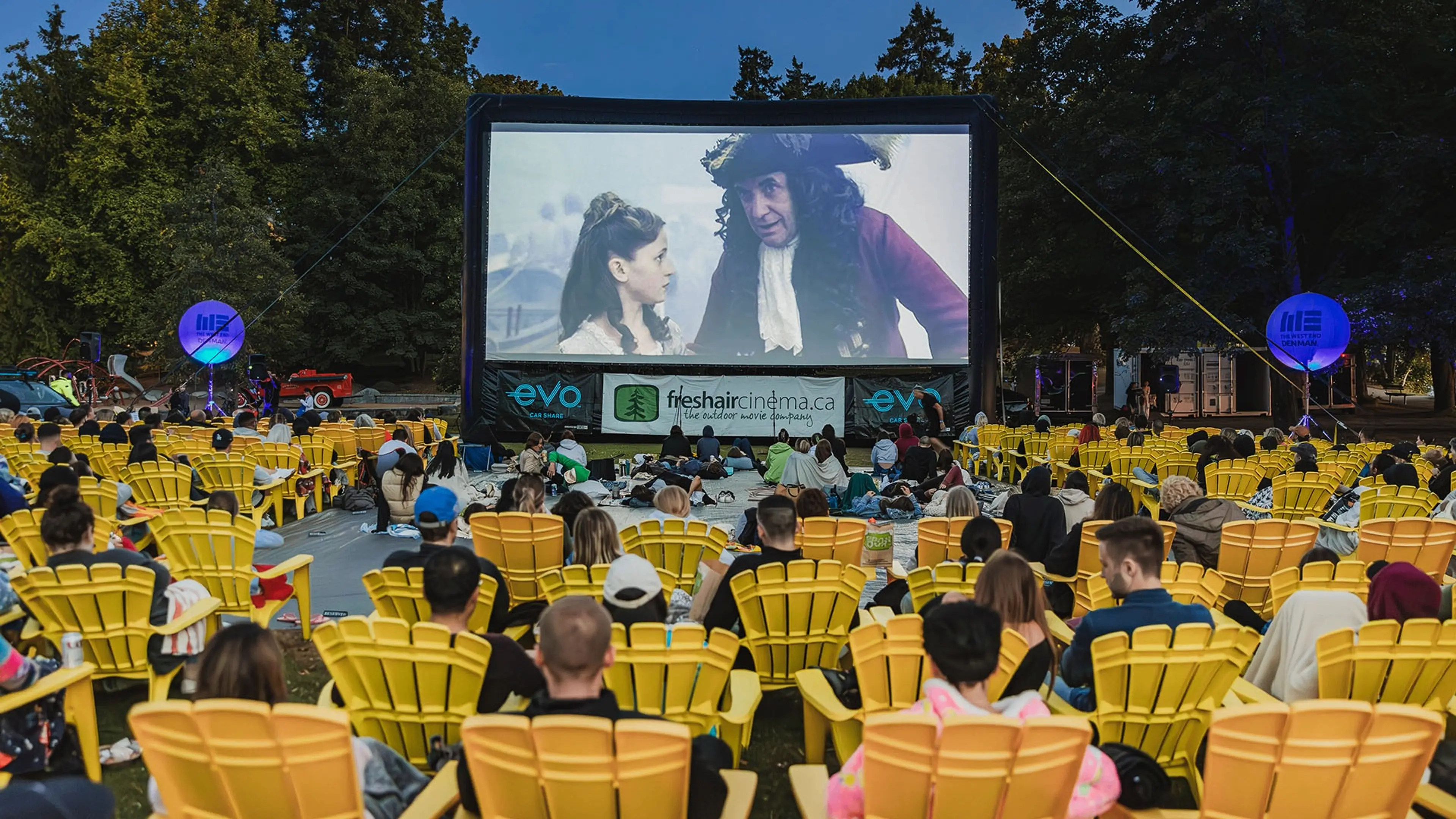 Outdoor movie screening with people seated in yellow chairs watching a film on a large screen surrounded by trees.