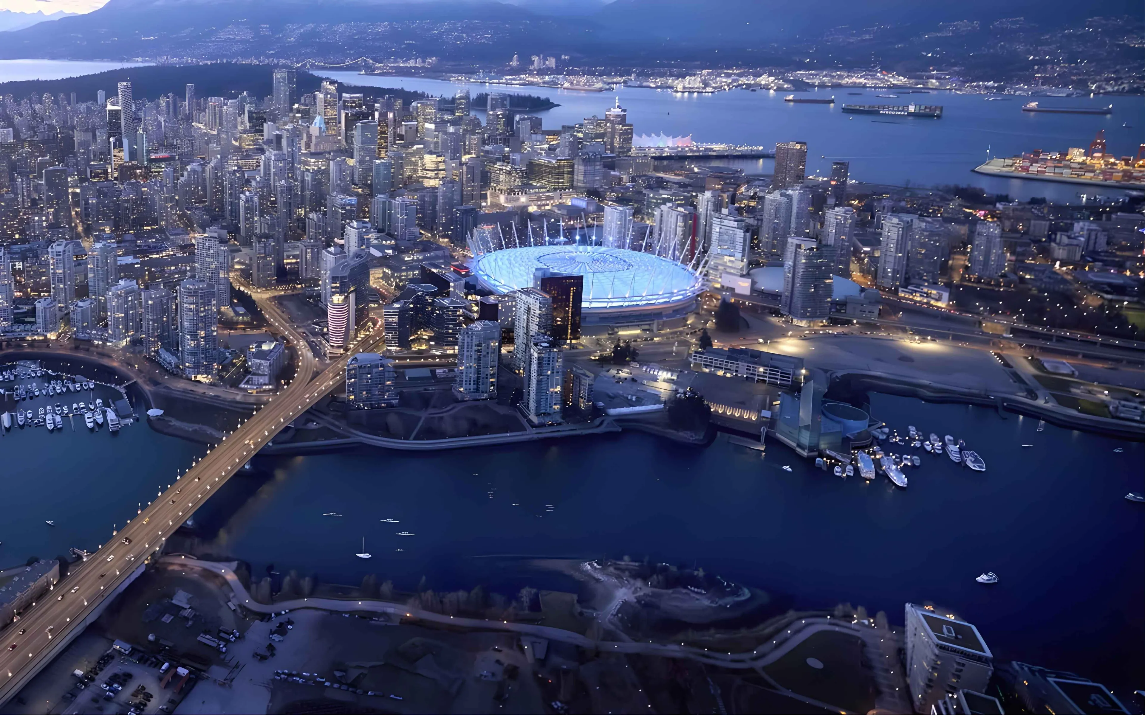 Aerial view of a Vancouver at dusk with a brightly lit stadium near the waterfront and a bridge crossing over the water.
