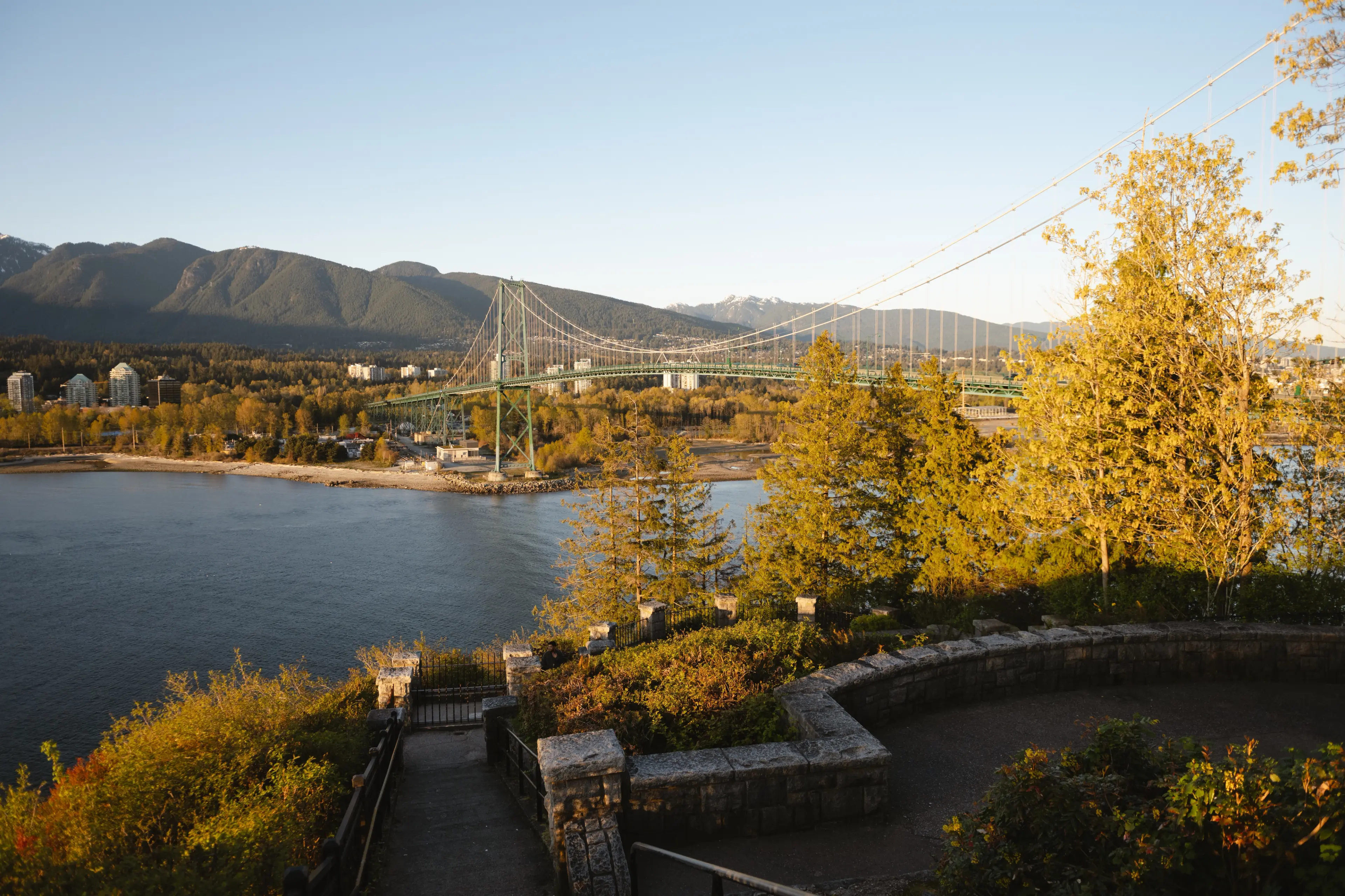 Suspension bridge over water with mountains and trees in autumn colors under clear sky.